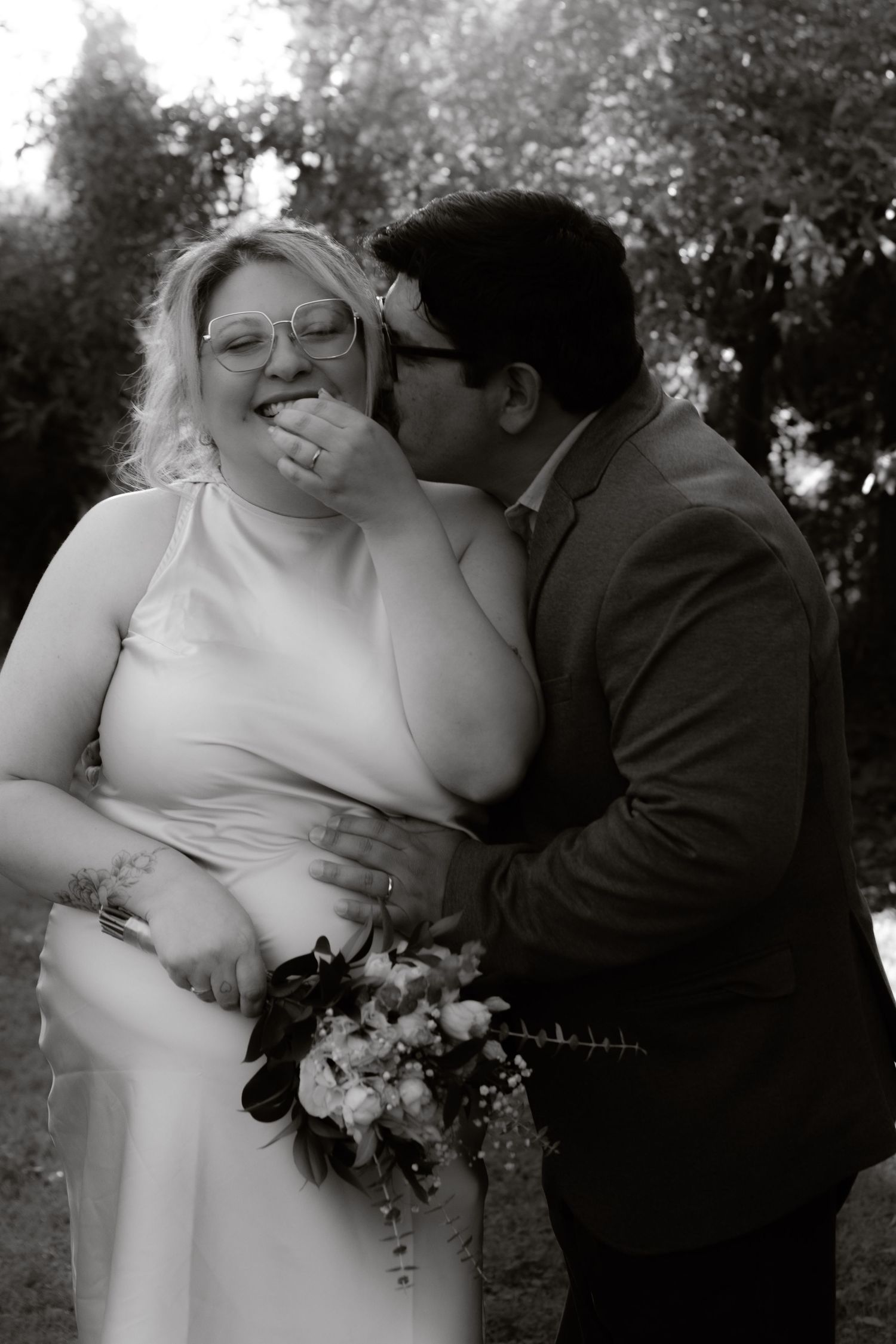 Black and white wedding photo of groom kissing laughing bride on cheek in garden setting.