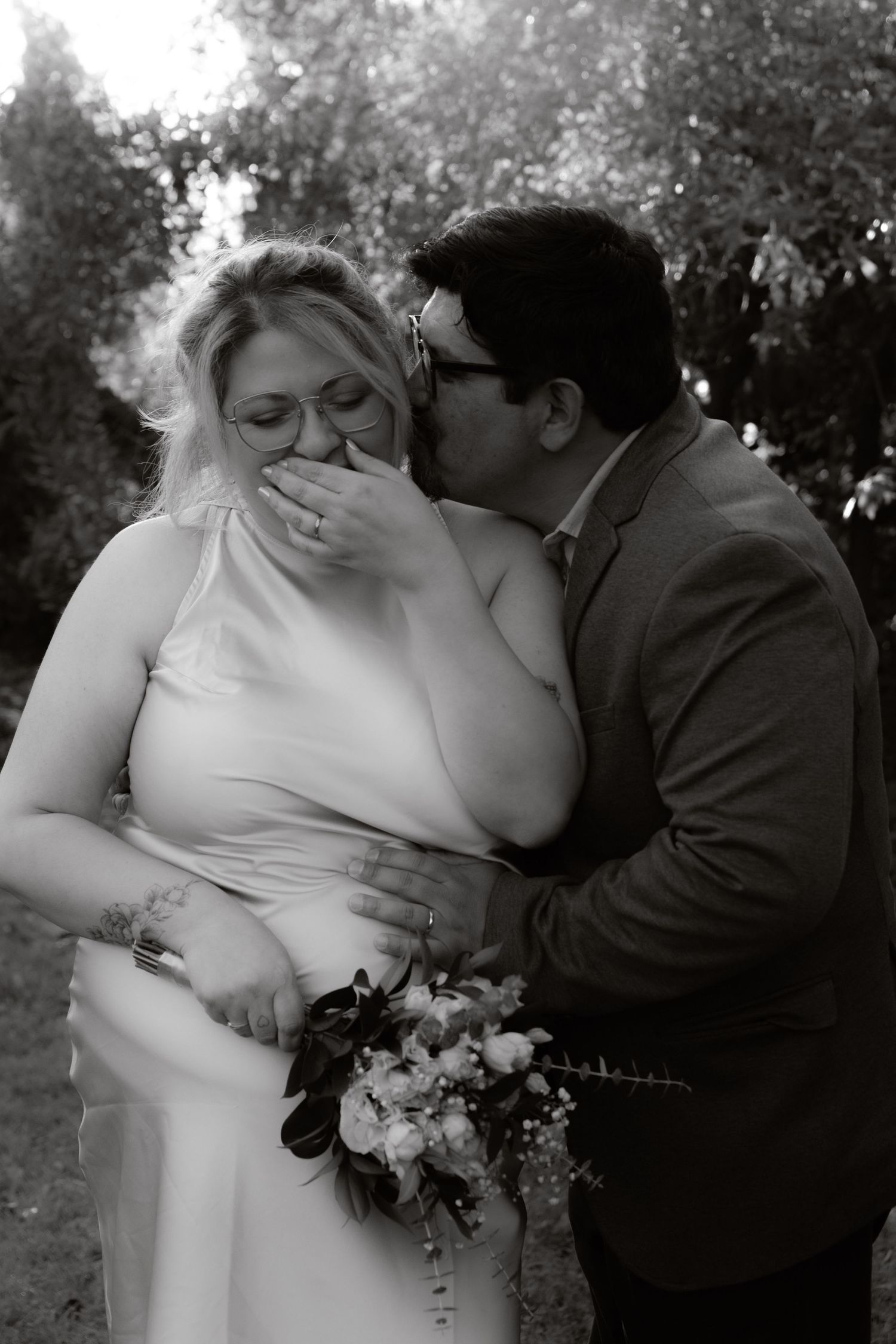 Black and white photo of groom kissing bride's cheek as she covers her smile with her hand.