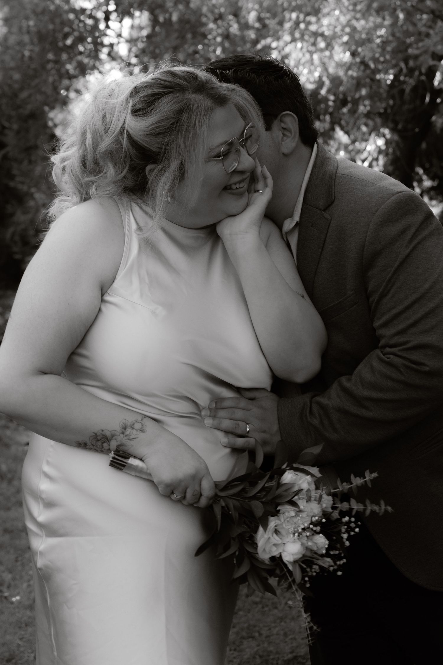 Black and white intimate wedding portrait of groom whispering to smiling bride in garden.