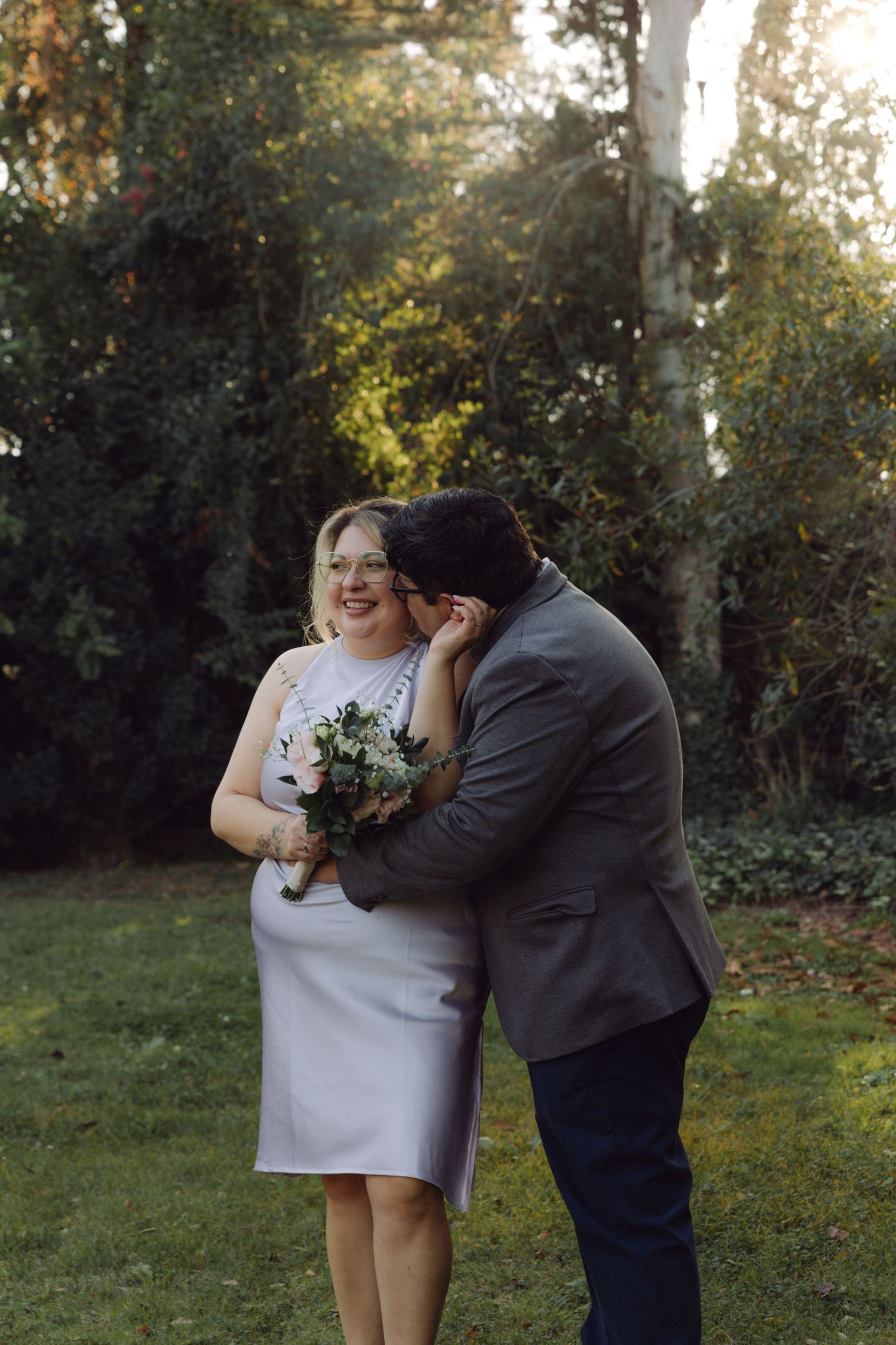 Bride in white dress laughing as groom leans in to kiss her cheek in sunny garden.