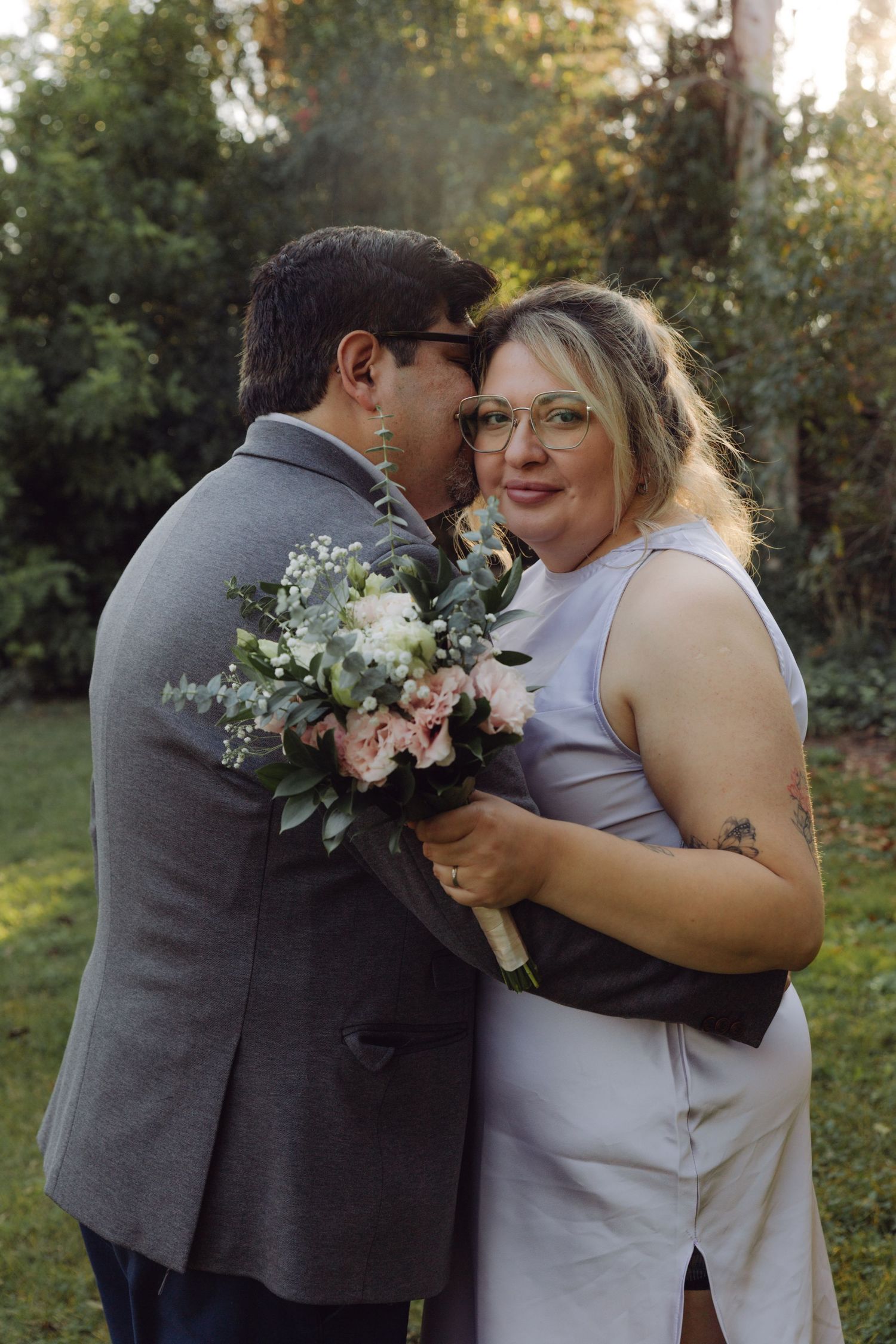 Groom in gray suit kissing bride's temple as she smiles holding a lush floral bouquet outdoors.