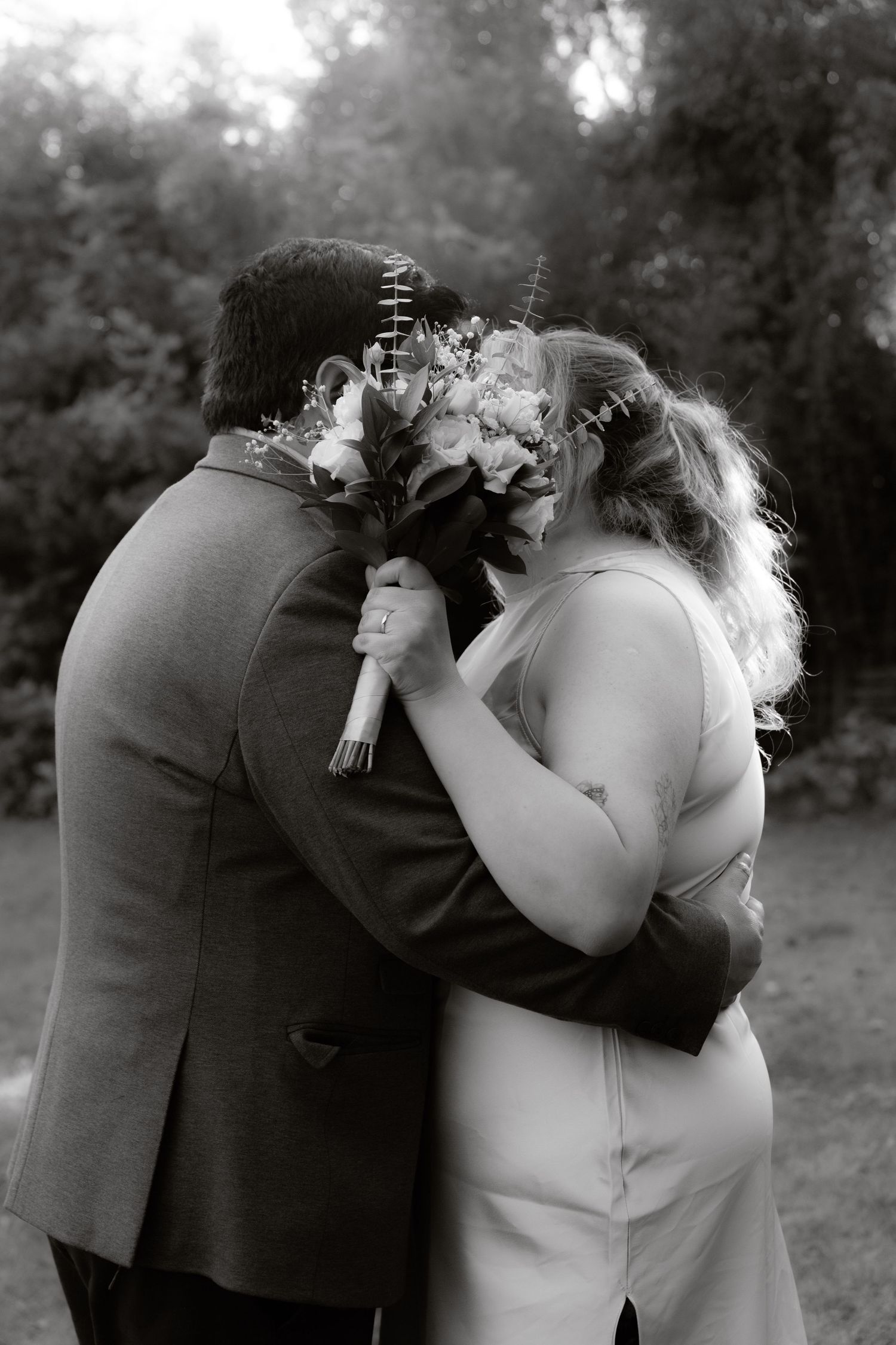 Black and white photo of newlyweds embracing from behind, bride holding bouquet in garden.