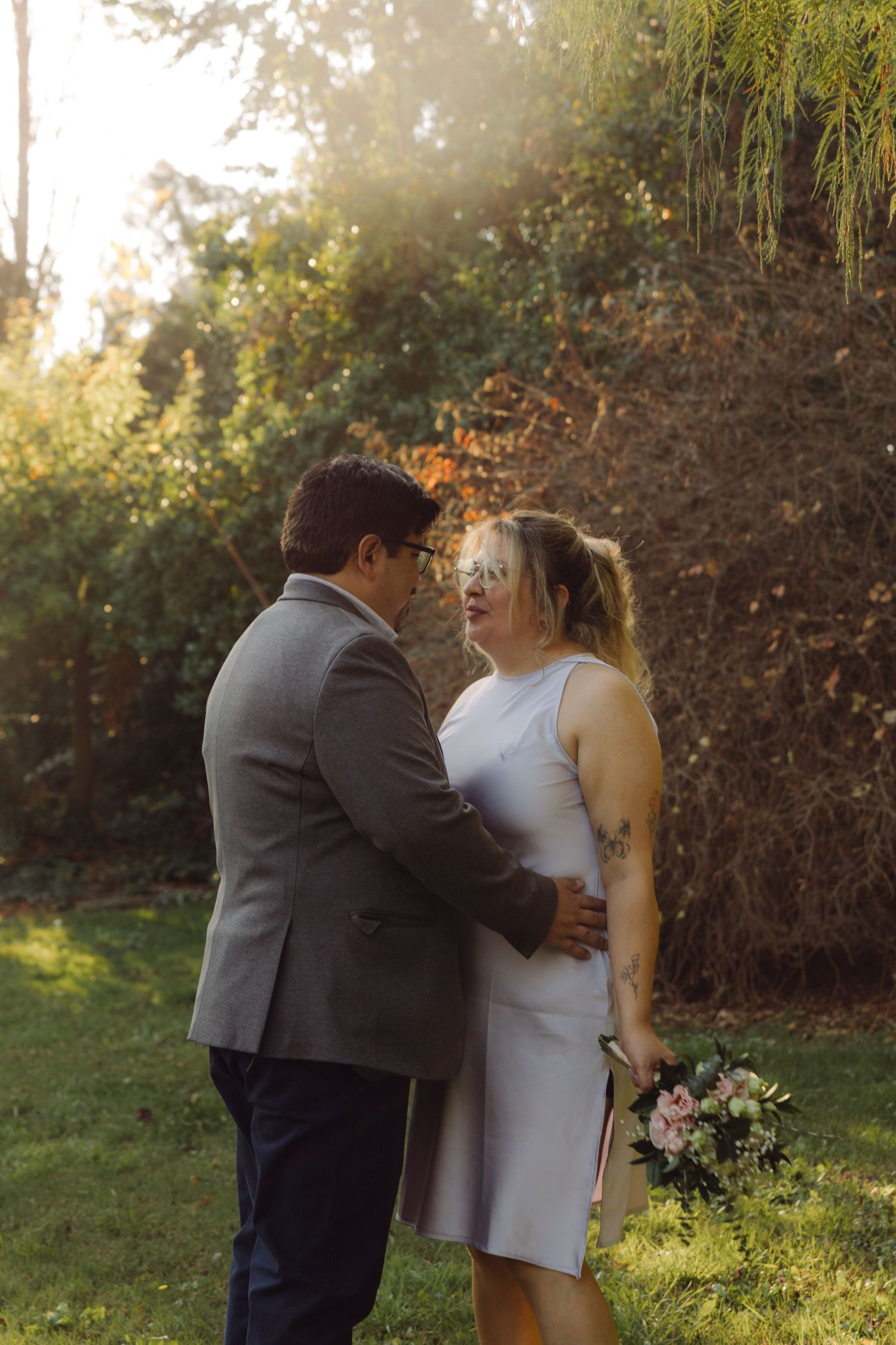 Couple embracing in golden sunlight during outdoor wedding ceremony, bride holding pink bouquet in white dress.