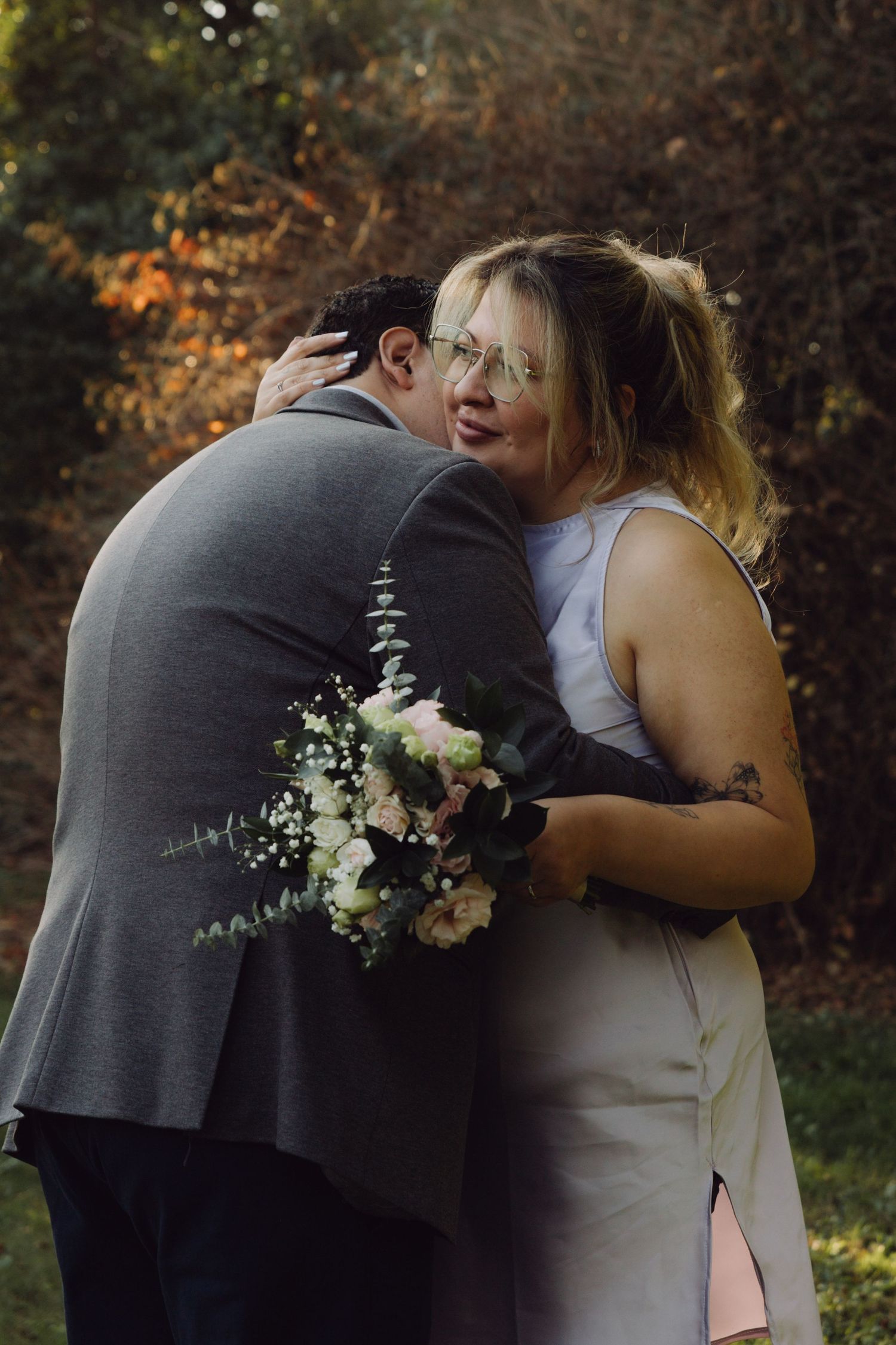 Newlyweds sharing a warm hug outdoors, bride holding floral bouquet with eucalyptus greenery at garden wedding.