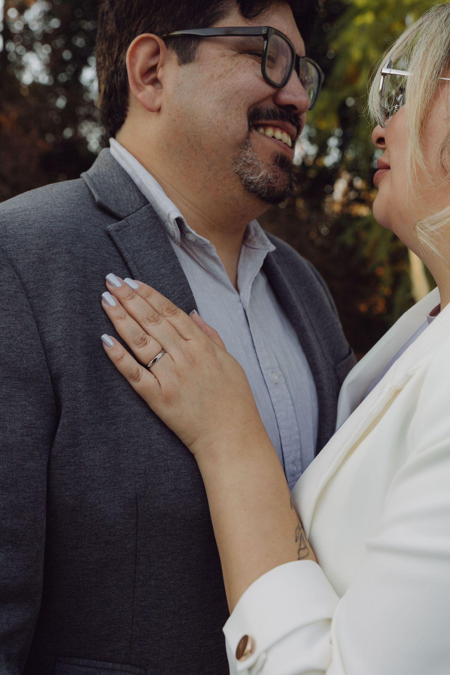 Close-up of smiling groom in gray suit with bride's hand on his chest showing wedding ring at outdoor ceremony.