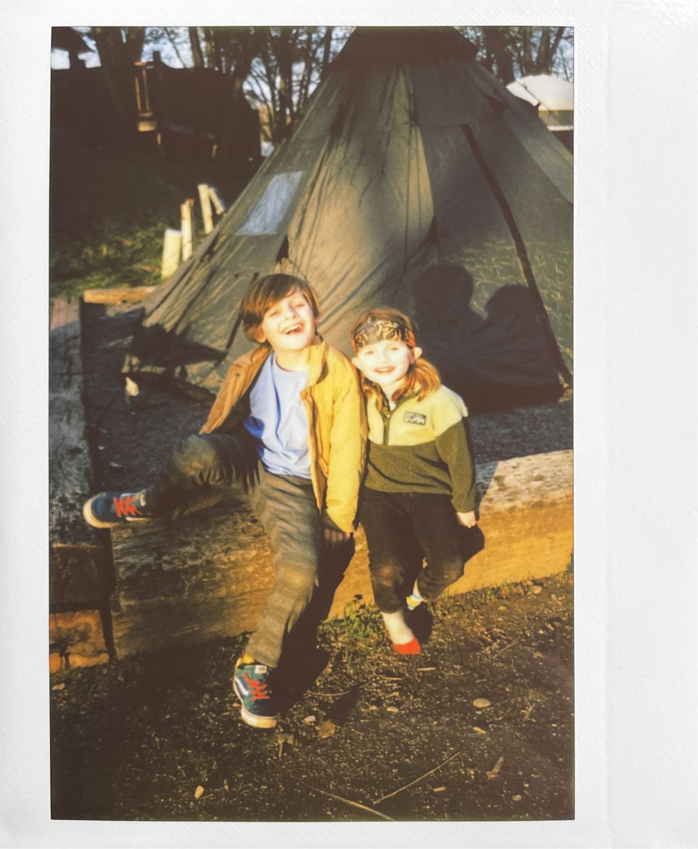 Two children in casual clothing stand together in front of a tent while camping outdoors.