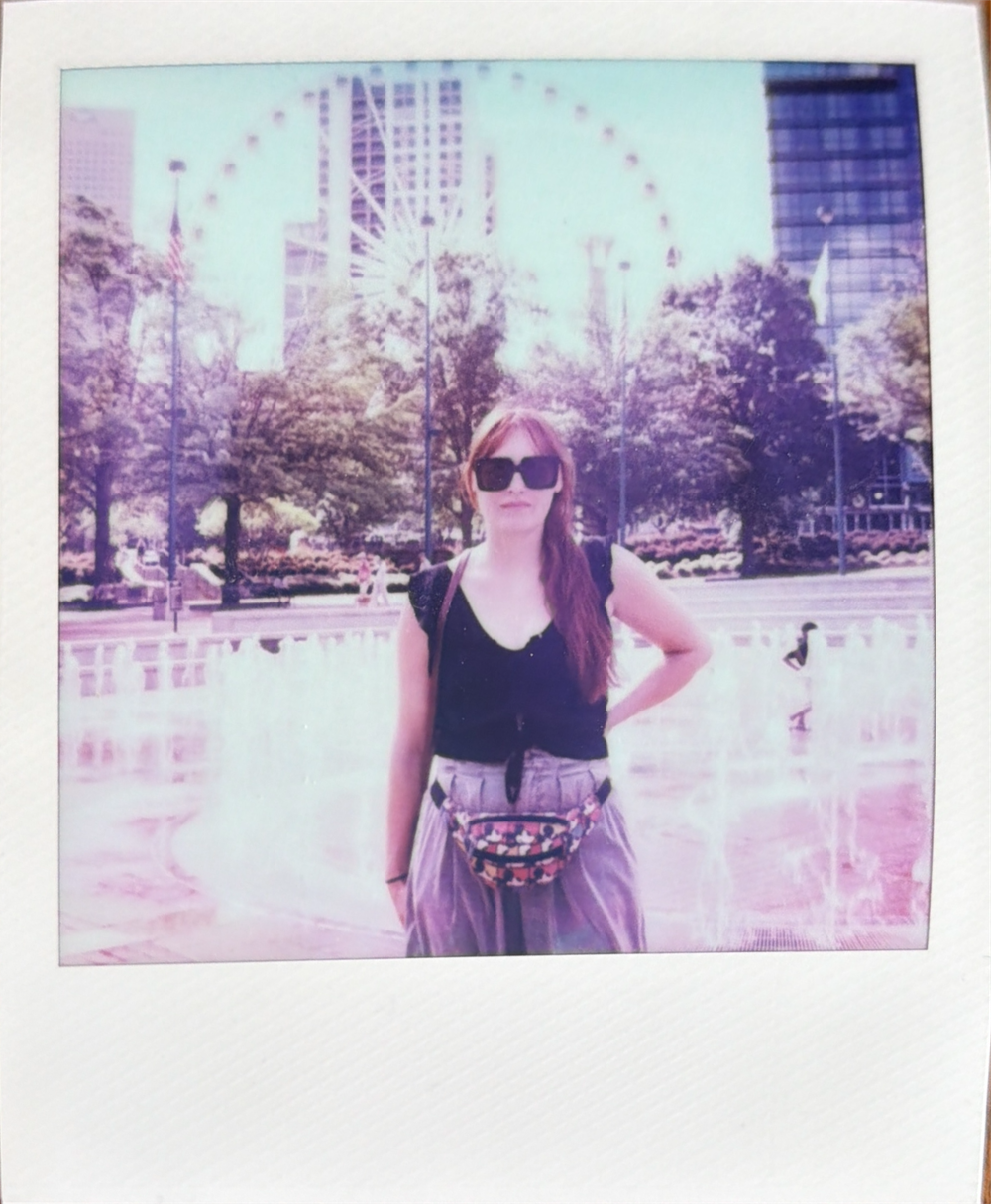 A person in sunglasses and black top poses in front of a Ferris wheel on a sunny day in an urban park.