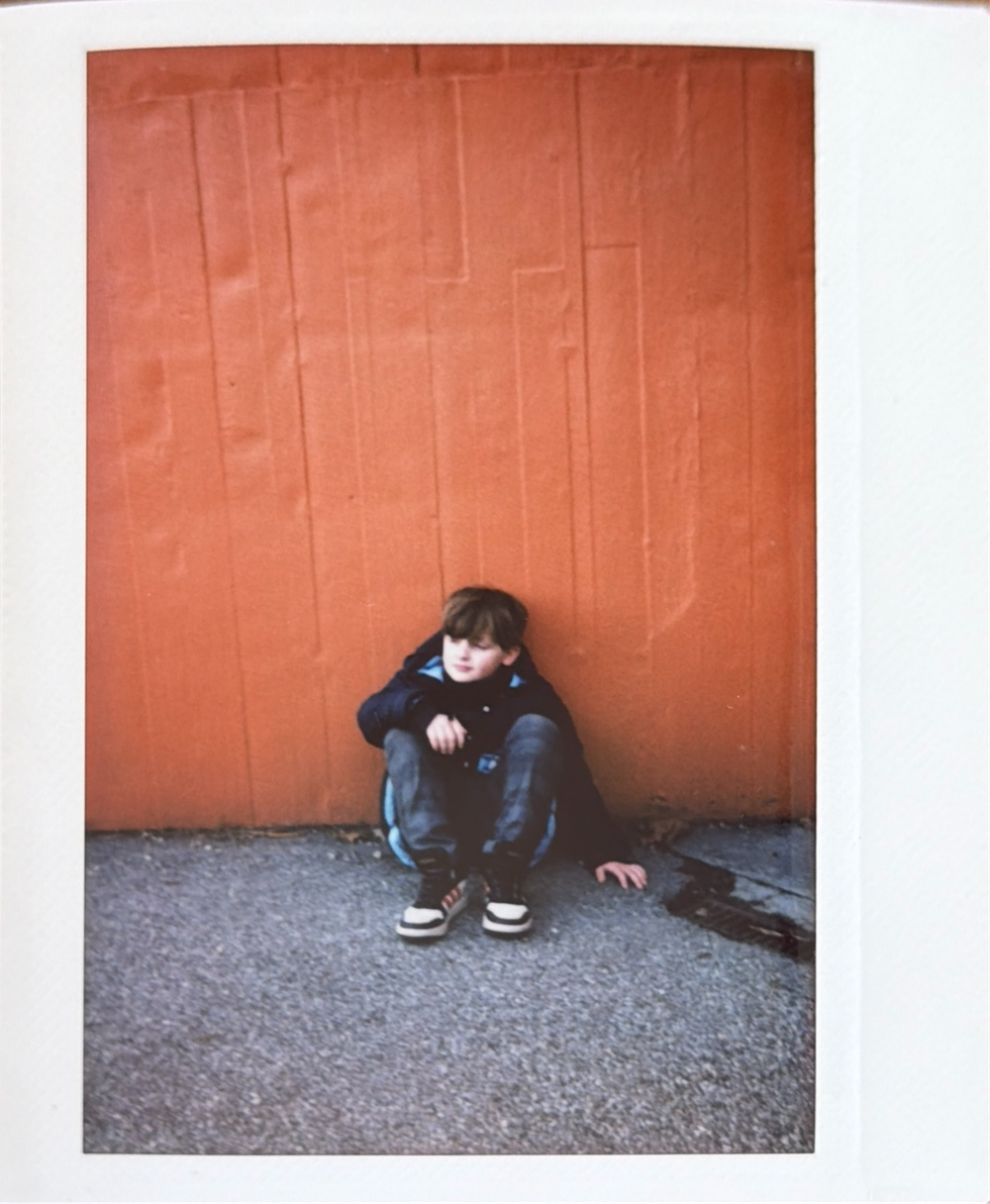 A person in dark clothing sits curled up against a terracotta-colored wall on a gray concrete floor.