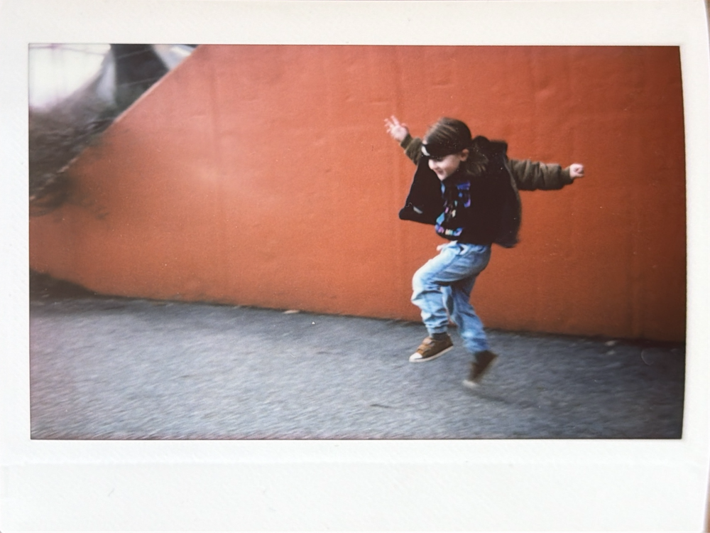 Person in blue jeans and black shoes performs a dynamic dance move against an orange wall background.