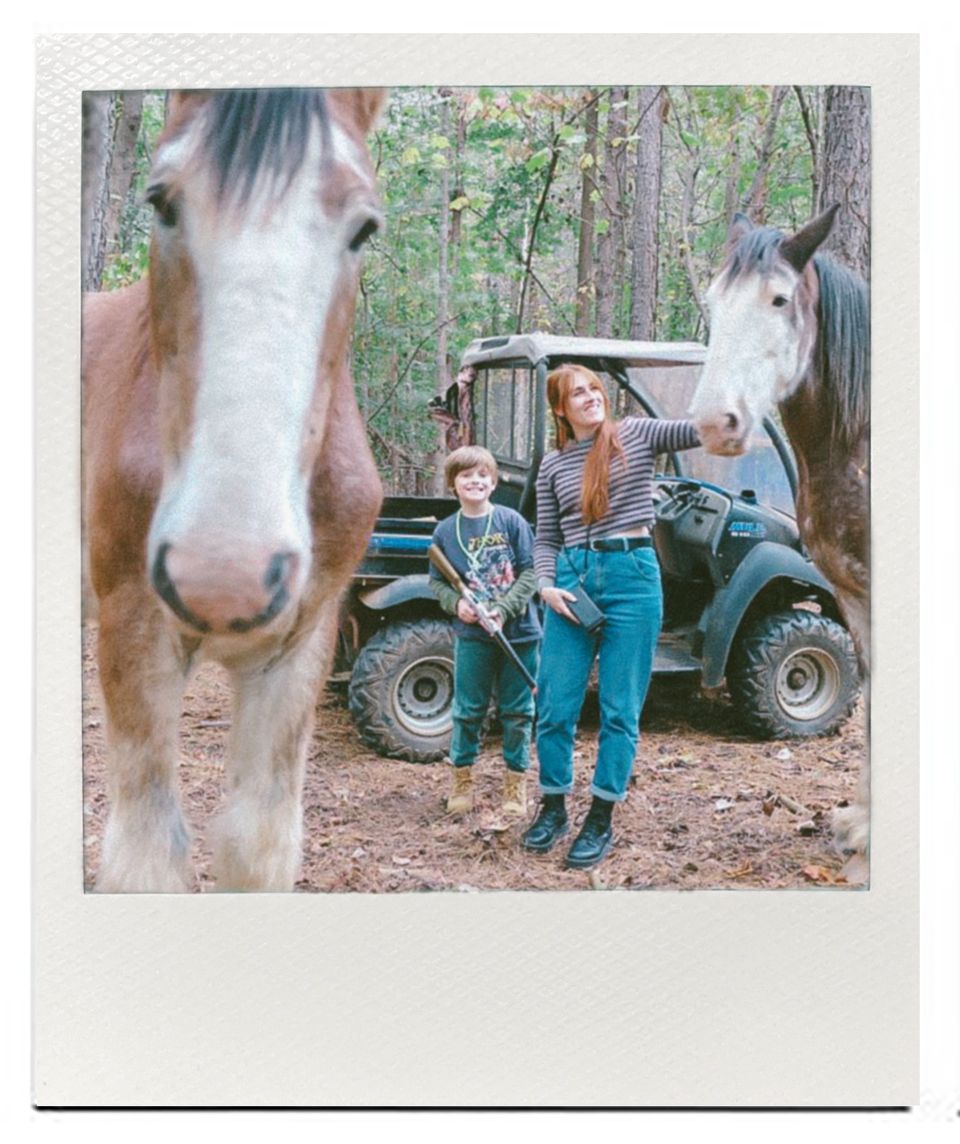 A young child in overalls stands next to a draft horse and vintage tractor in a wooded setting.