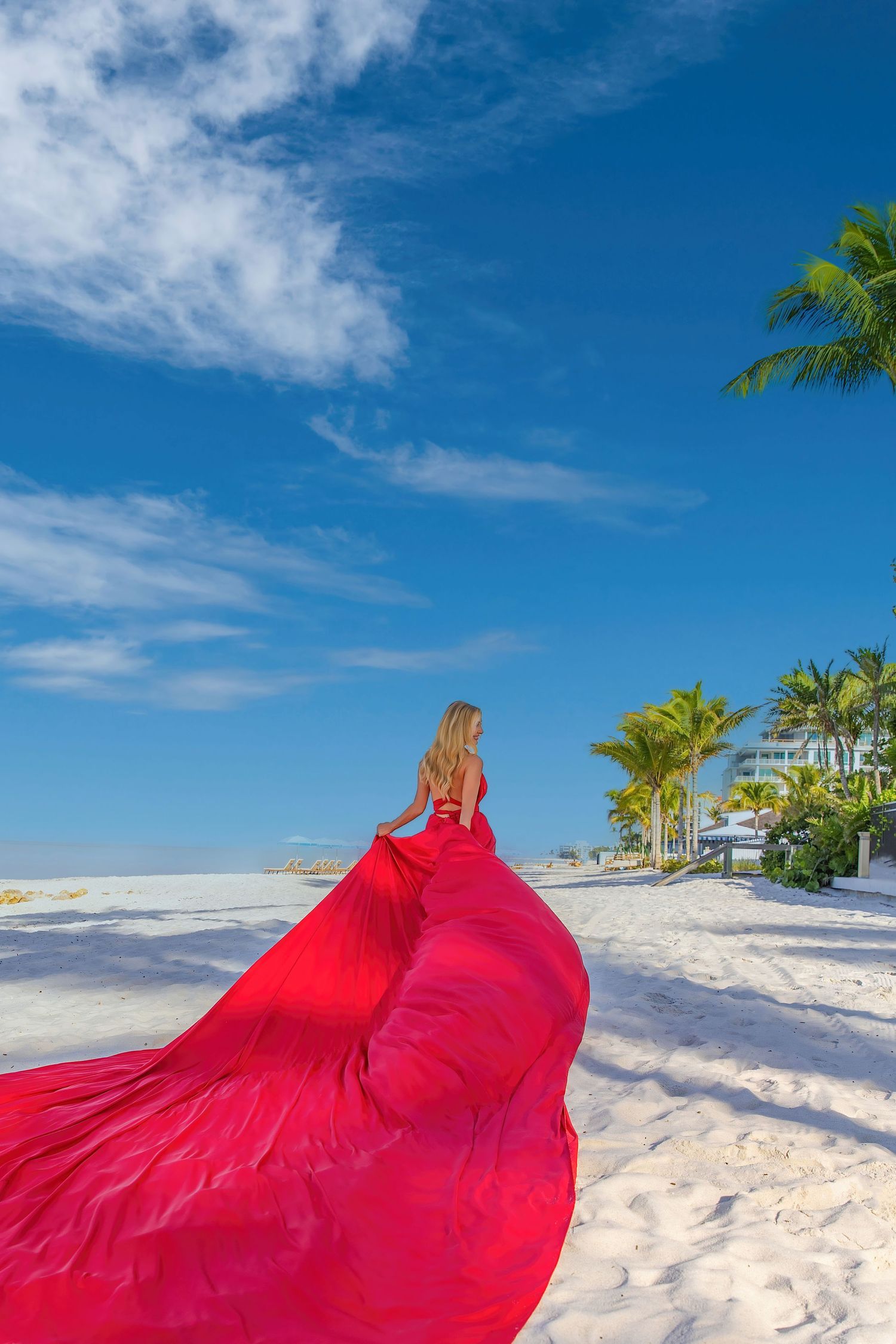 A woman in a flowing red gown with long train poses elegantly between dramatic classical columns with sunlight streaming in. Flying Dress