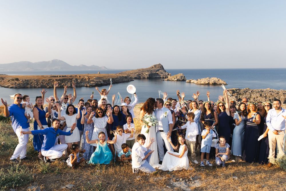 Large group of wedding guests in white and blue attire pose together on a scenic coastal cliff at sunset.