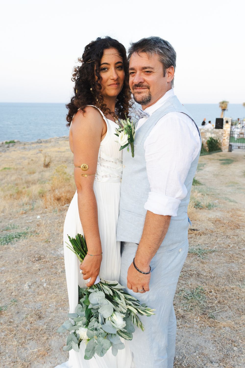 Couple in white wedding attire pose together on coastal clifftop during sunset ceremony.