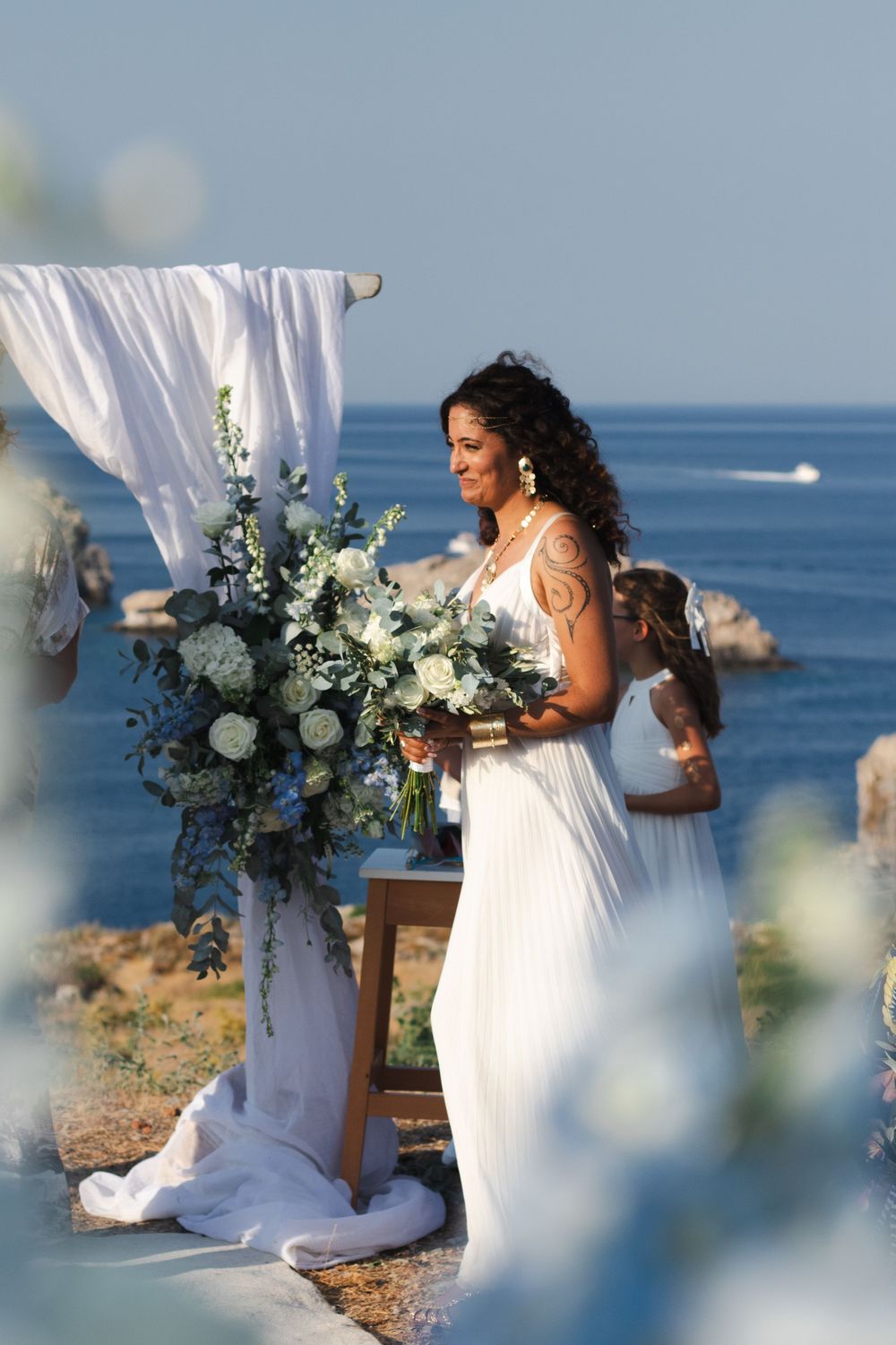 Seaside wedding setup with white flowing fabric and blue floral arrangements against ocean backdrop.