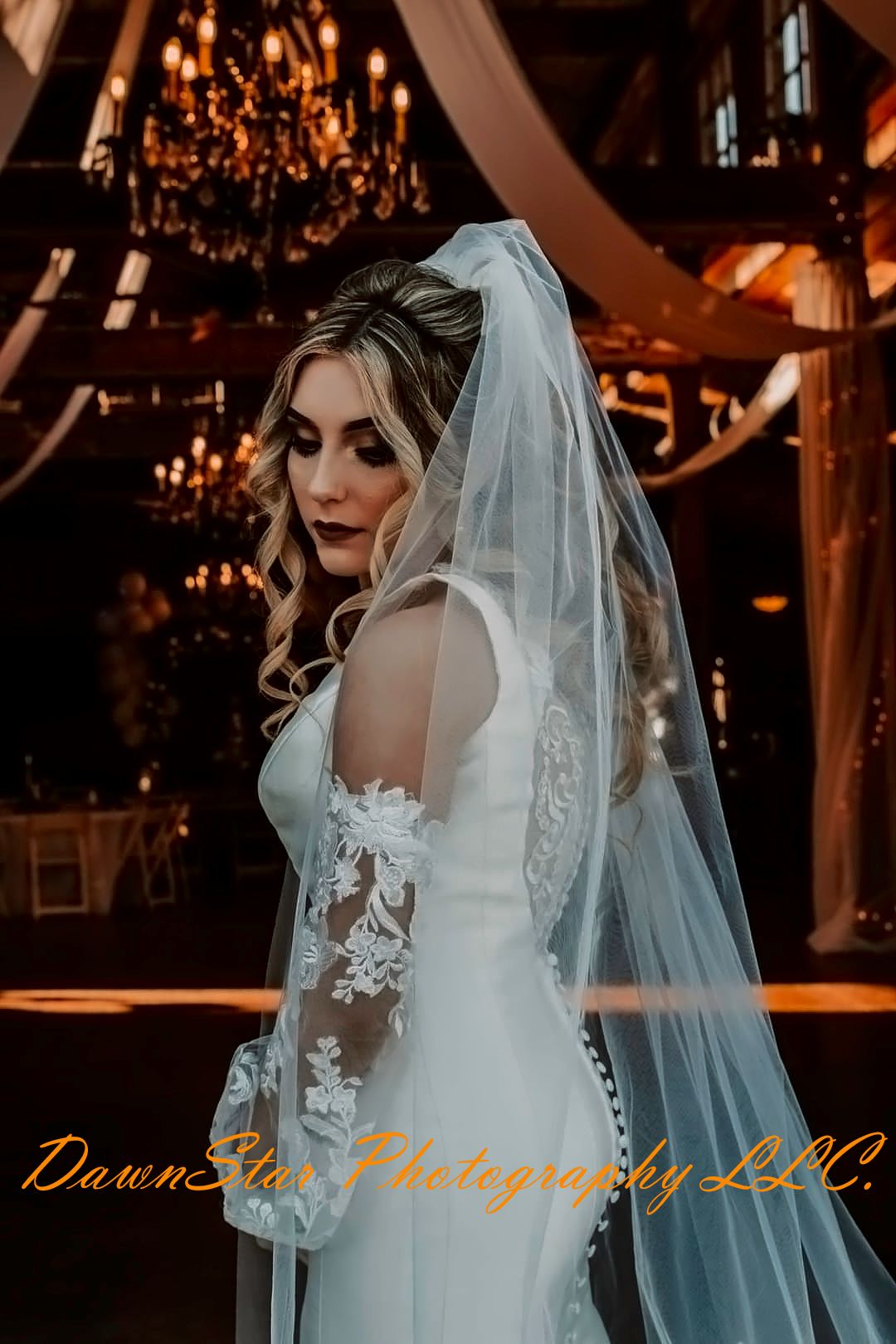 Elegant bride in white lace wedding gown and flowing veil poses under crystal chandeliers in romantic venue setting.