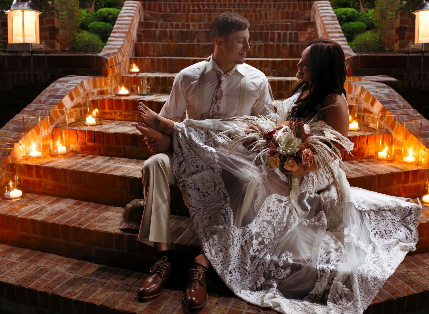 A dramatic wedding dress train flows across rustic brick steps illuminated by romantic candlelight at dusk.