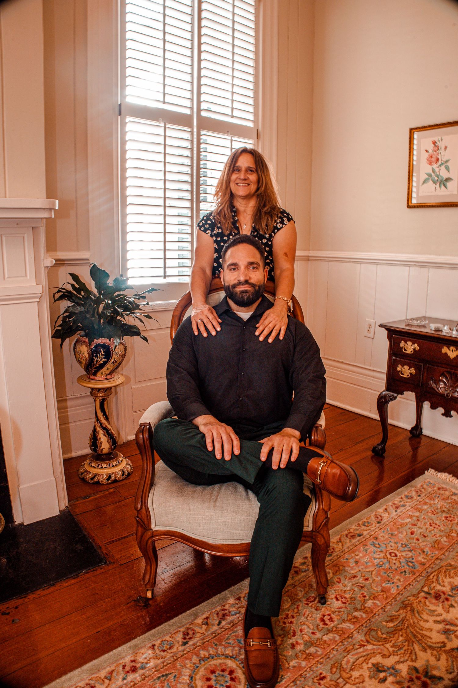 A couple poses in an elegant room, man seated in ornate chair, woman standing behind with hands on his shoulders.