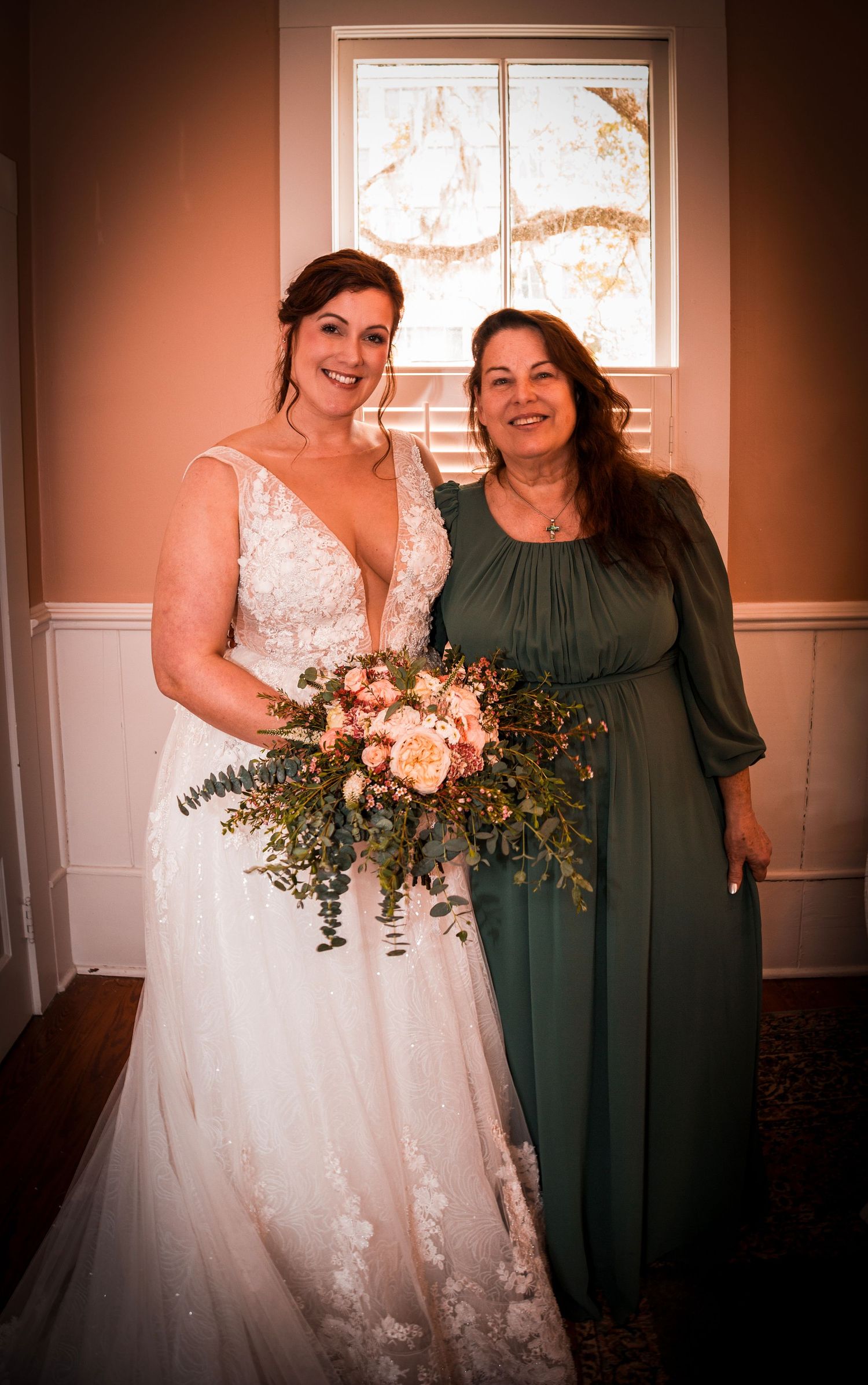 Bride in white lace gown holding pink floral bouquet poses with woman in green dress near window.