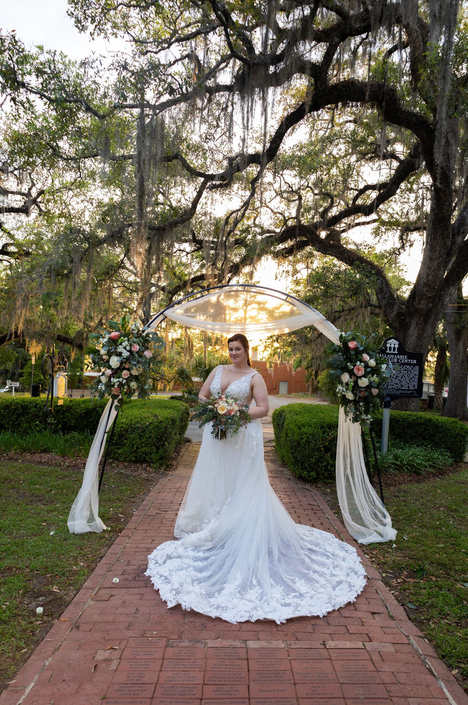 Bride in lace wedding gown stands under floral arch on brick path, framed by Spanish moss-draped oak trees at sunset.