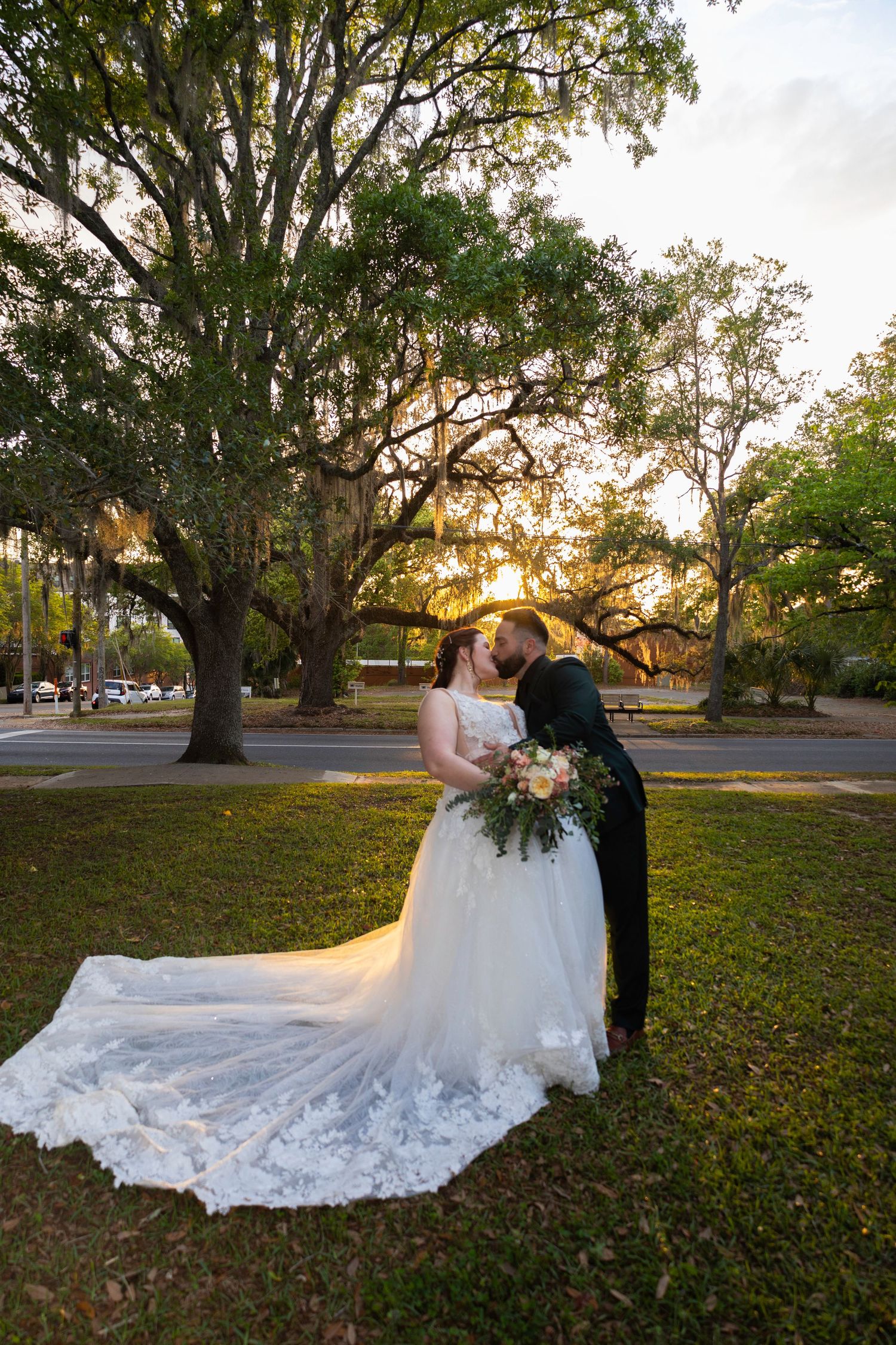 Bride and groom share a romantic kiss at sunset beneath sprawling oak trees, her lace gown trailing on lush green grass.