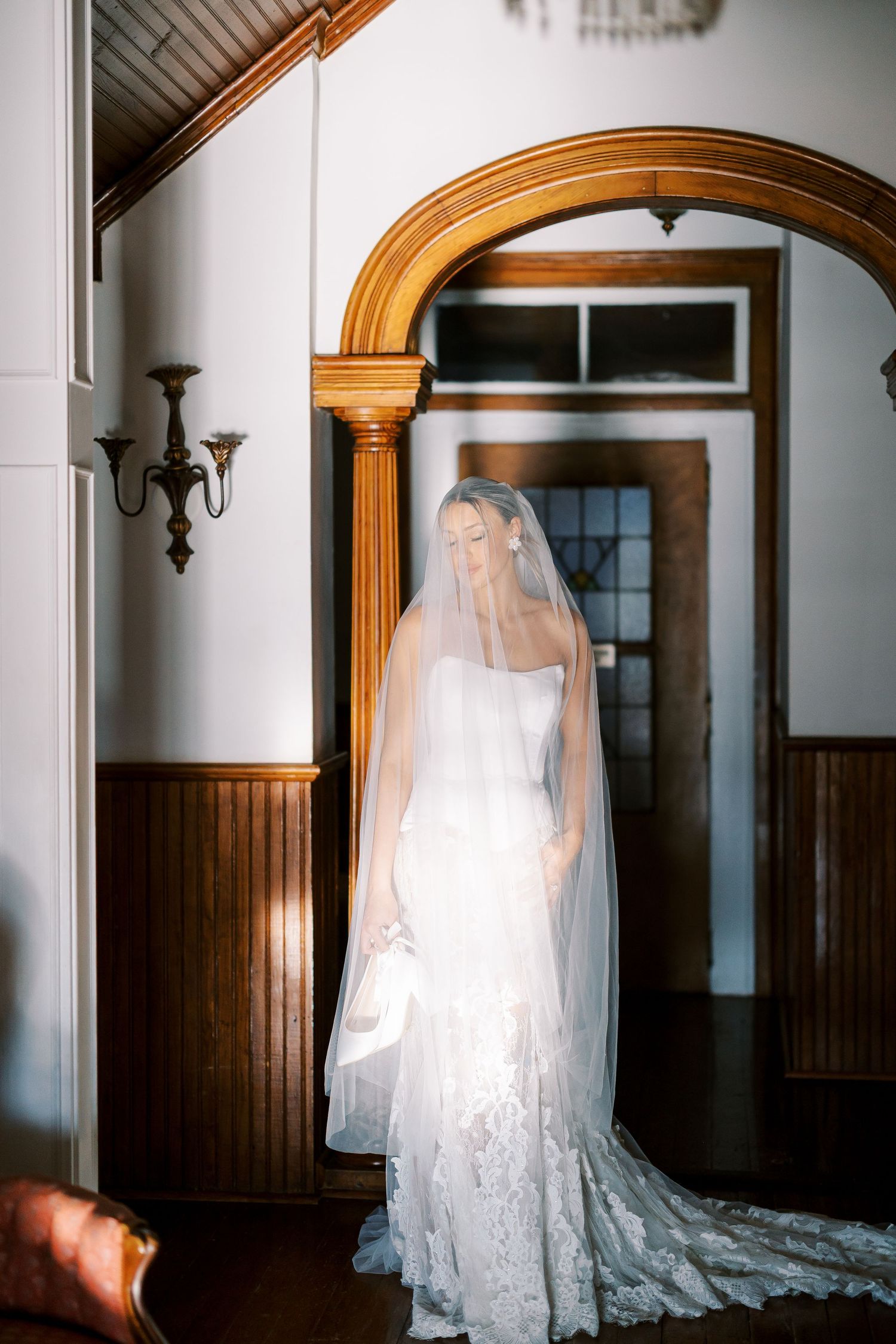 A bride in a stunning lace wedding gown and long veil stands elegantly in front of a wooden archway.