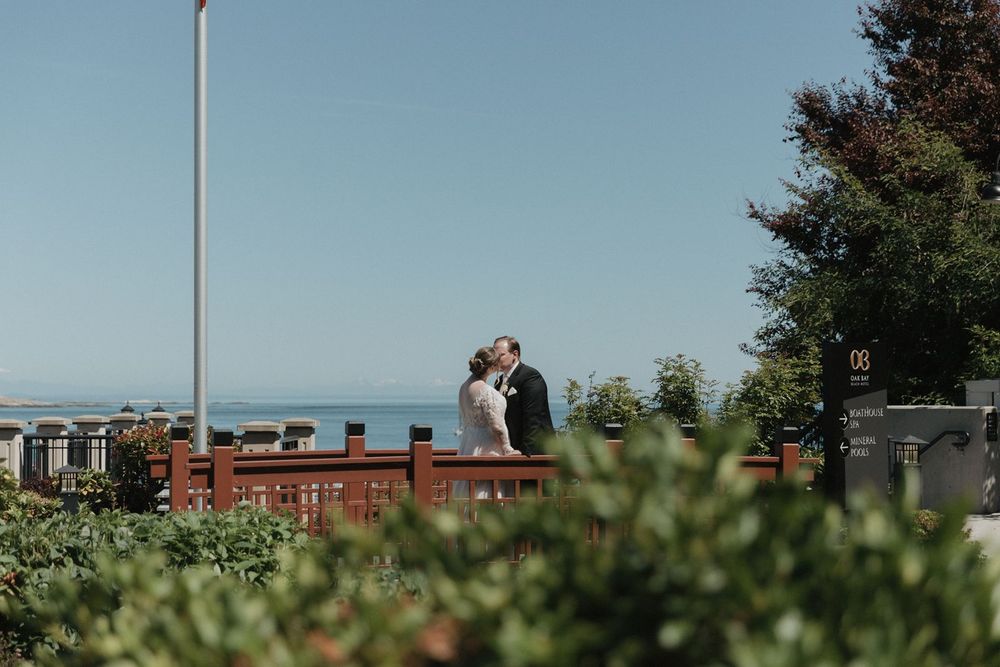 Bride and groom share a kiss on a scenic overlook with ocean views and lush greenery framing the moment.