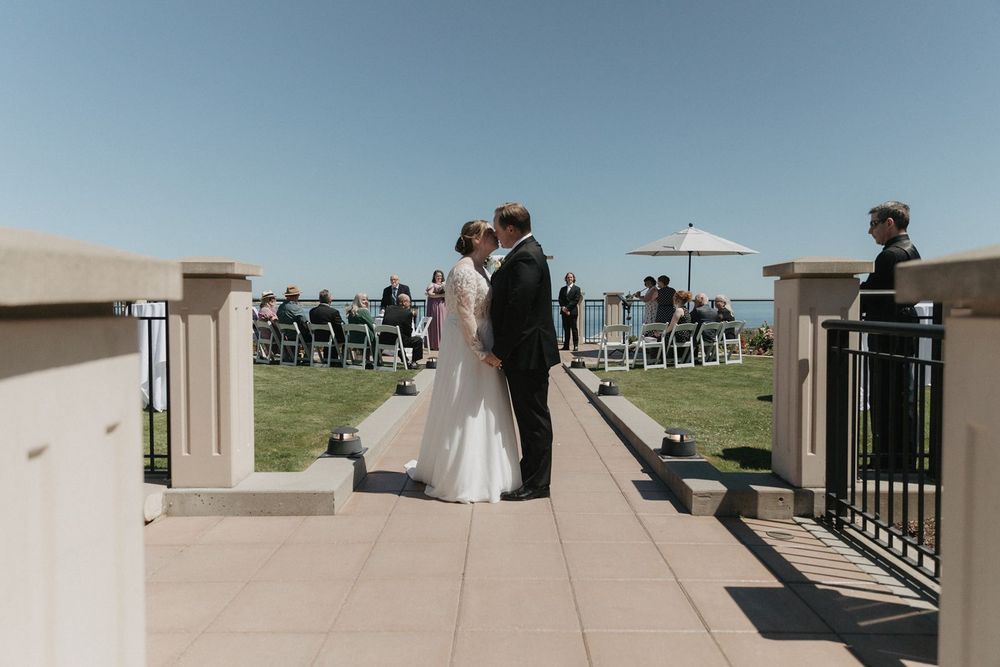 Bride and groom share first kiss at outdoor waterfront wedding ceremony with white chairs.