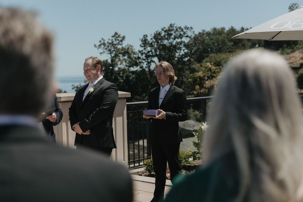 Groom and officiant stand at outdoor wedding ceremony overlooking a scenic waterfront venue.
