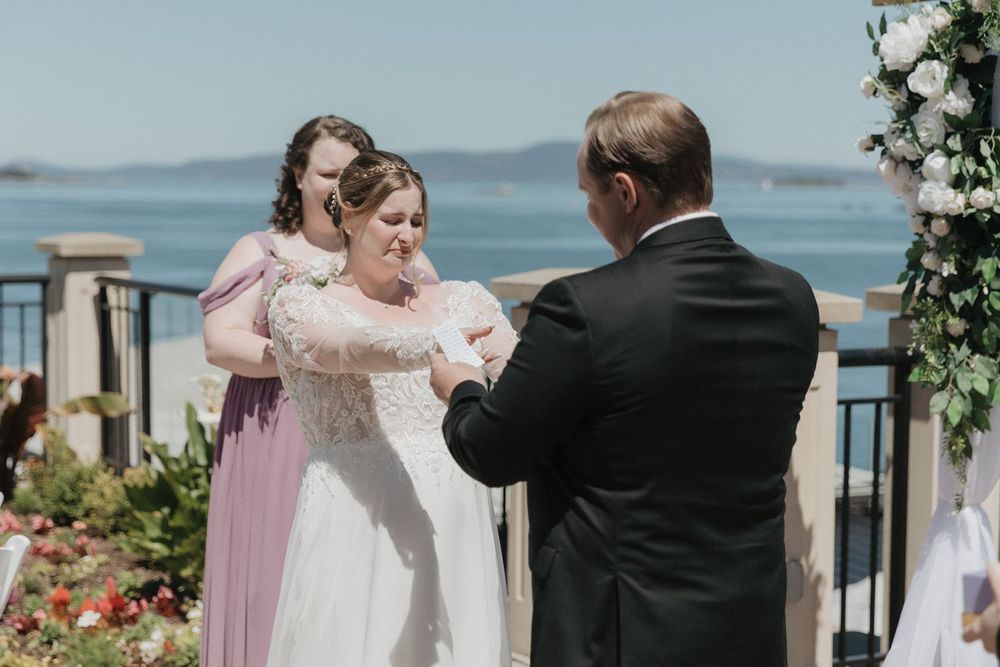 Bride receives ring from groom during outdoor waterfront wedding ceremony with bridesmaid nearby.