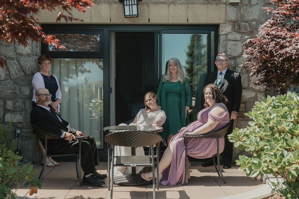 Wedding party group photo on a sunlit patio surrounded by lush greenery and red foliage.