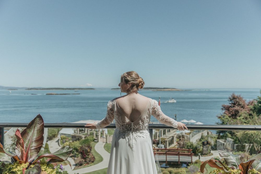 Bride in lace wedding dress stands on balcony overlooking serene ocean view on clear sunny day.