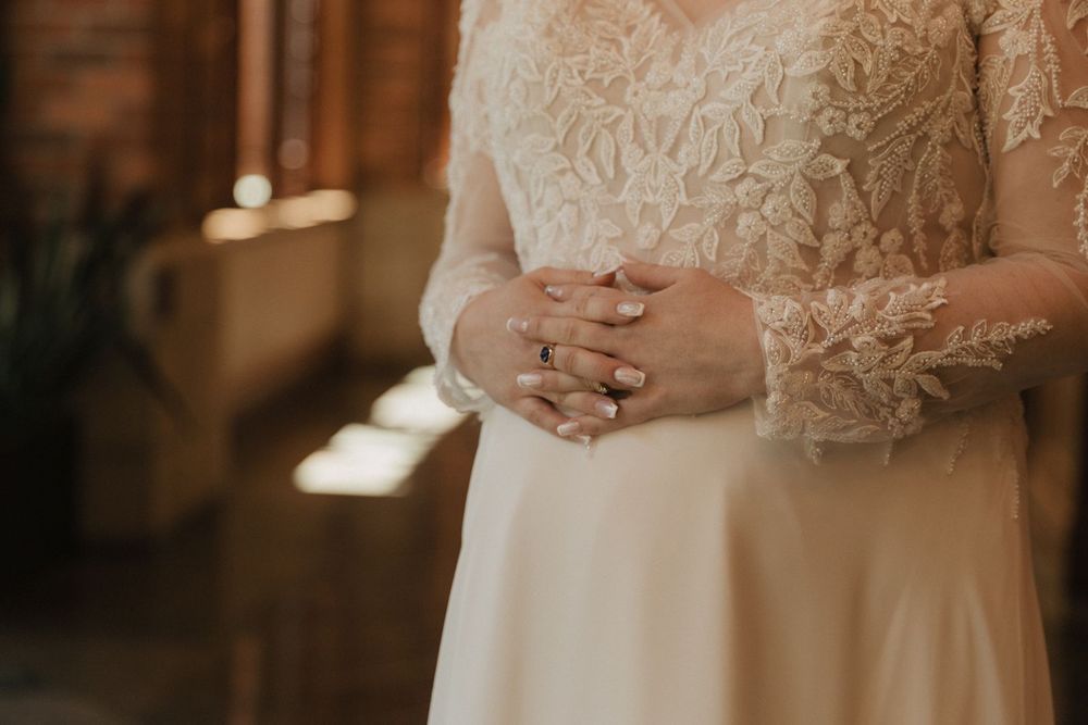 Close-up of bride's hands clasped together wearing lace long-sleeve wedding gown and ring.