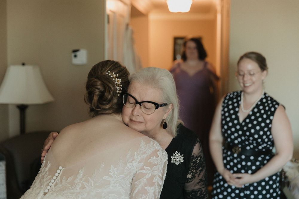 Elderly woman embracing bride in lace wedding dress while guests smile warmly in background.