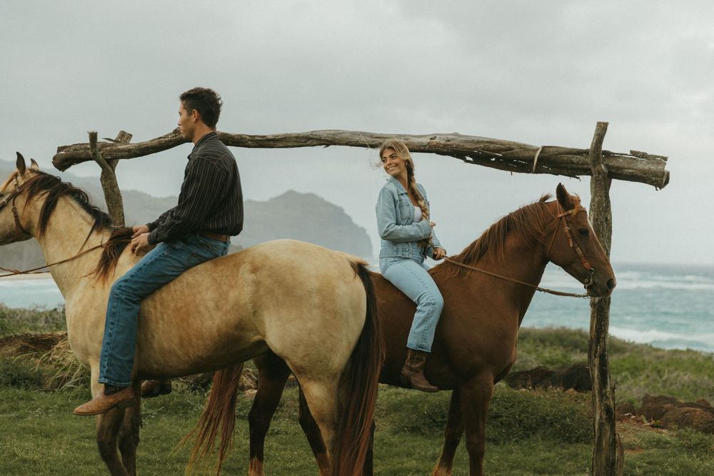 Horses and riders gathered on a green hillside with a rustic wooden fence and ocean backdrop.