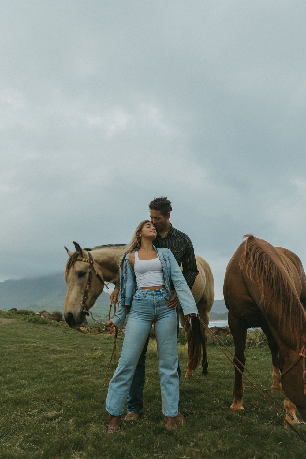 A couple stands with two horses in a grassy field against a cloudy sky and mountain backdrop.