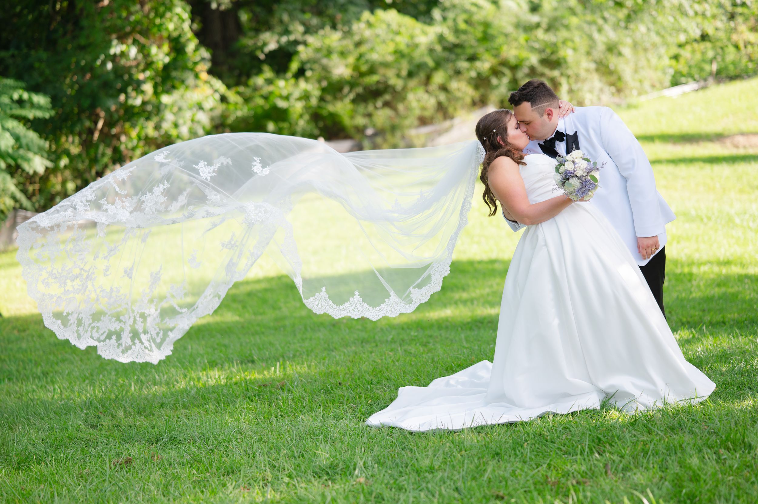 A flowing white wedding veil blows gracefully in the wind while newlyweds kiss on the lawn at Las Mañanitas in Brewster, NY .