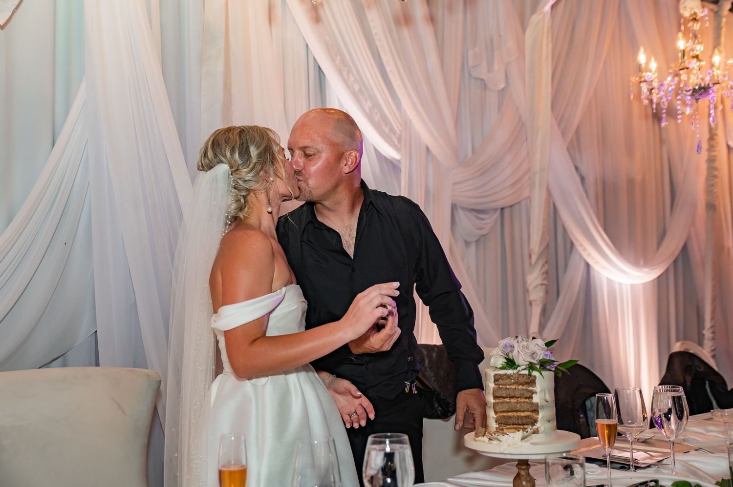 Couple dances on black and white checkered floor at wedding reception with purple-clad wedding party watching.