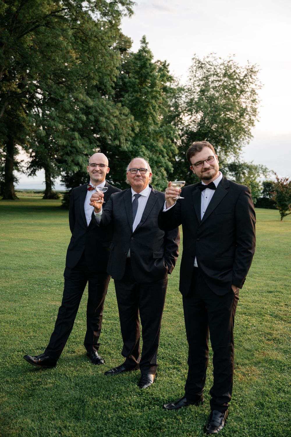 Groomsmen in black tuxedos pose together on a green lawn during an outdoor wedding.