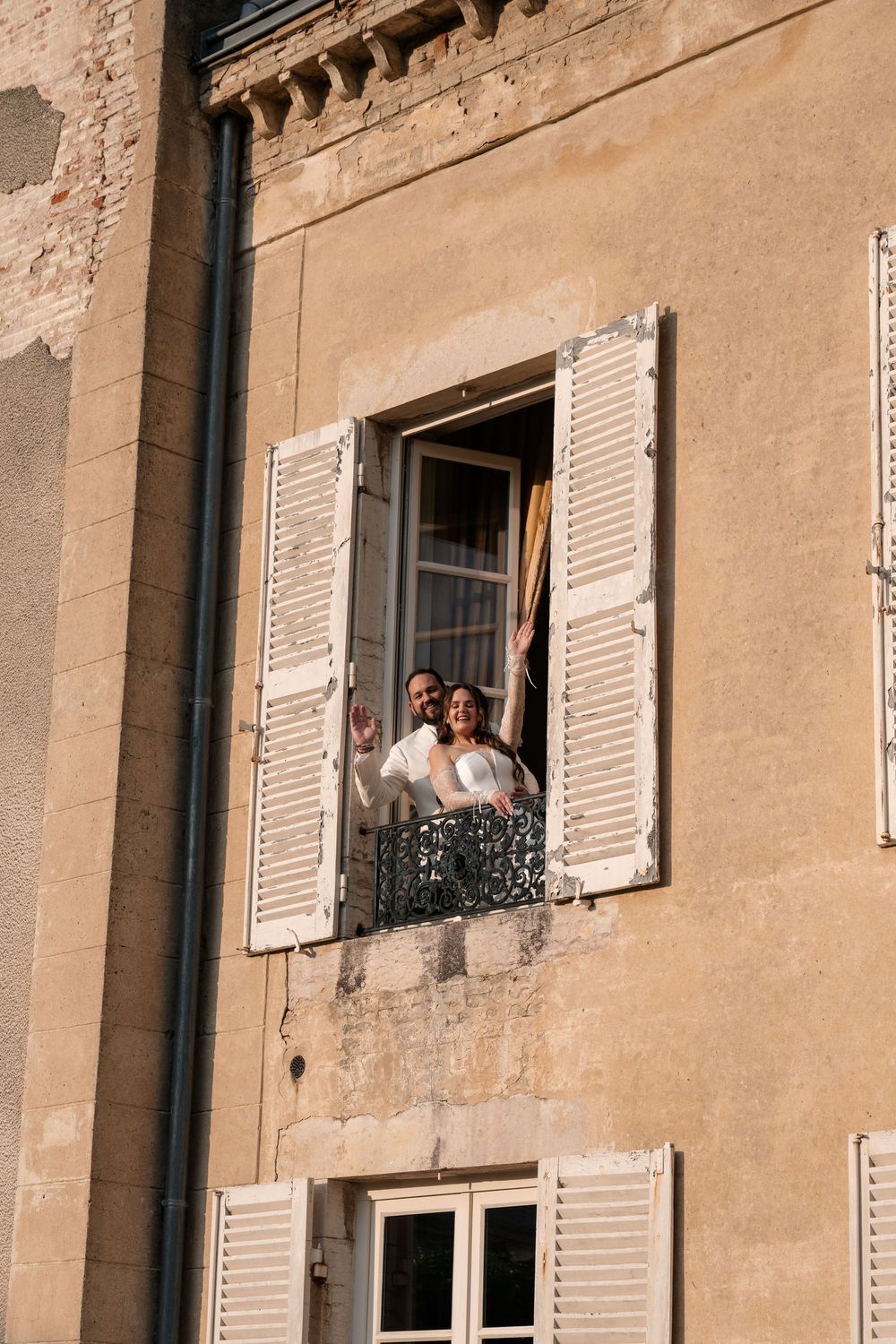 Two figures lean out of a rustic window with white shutters in a beige stone building.