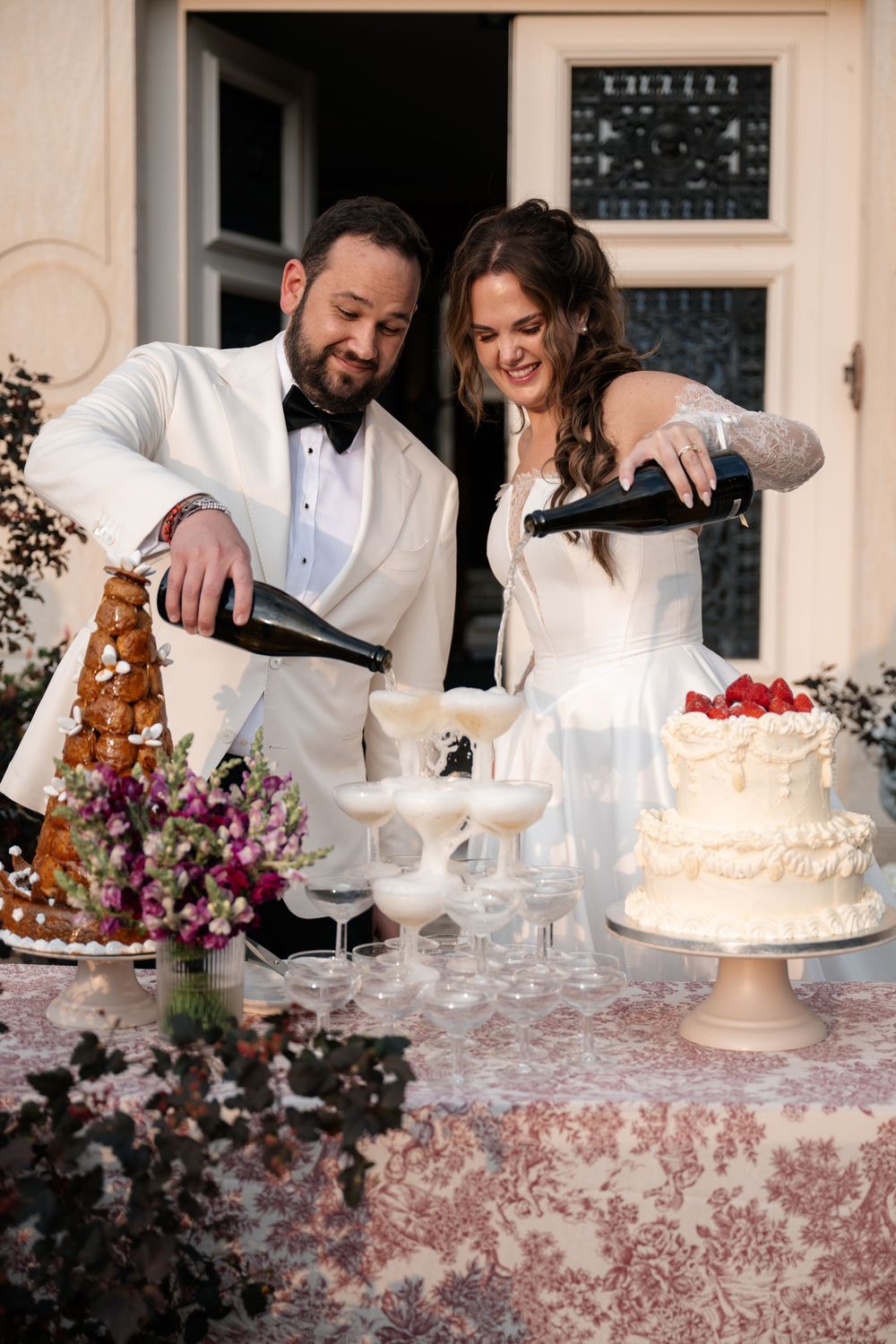 Wedding couple cutting and serving elegant white tiered cakes at an ornate dessert table.