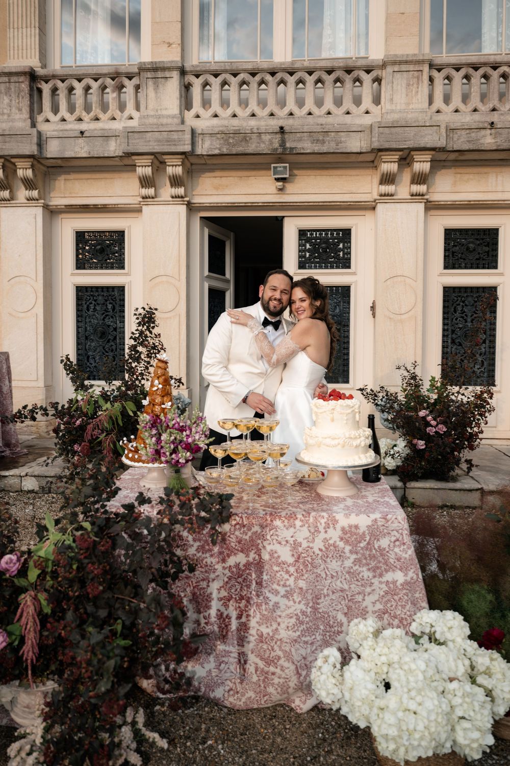 A romantic wedding couple sits on an elegant stone balcony surrounded by floral arrangements and white hydrangeas.