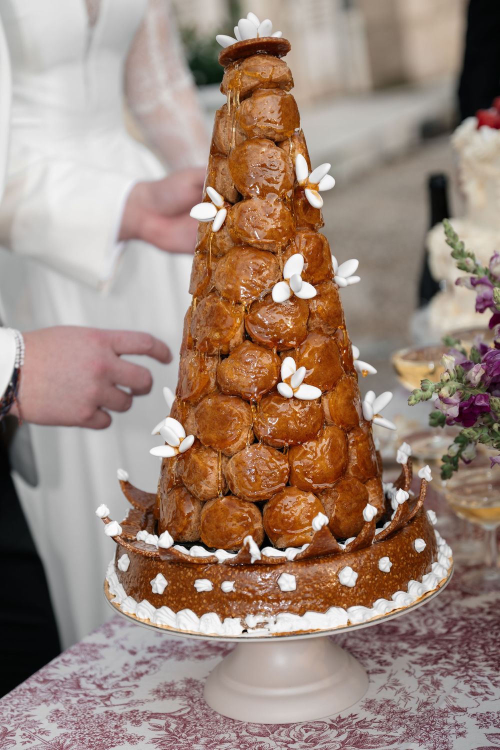 A traditional croquembouche wedding cake with caramel-glazed profiteroles and white floral decorations on a cake stand.