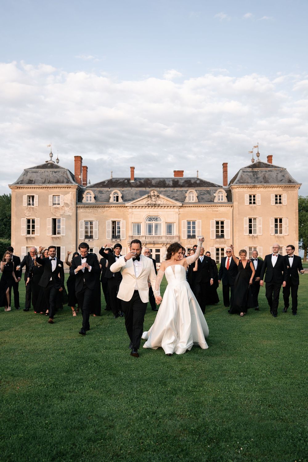 Wedding party poses on a grand lawn in front of a French chateau with bridesmaids in white and groomsmen in black.