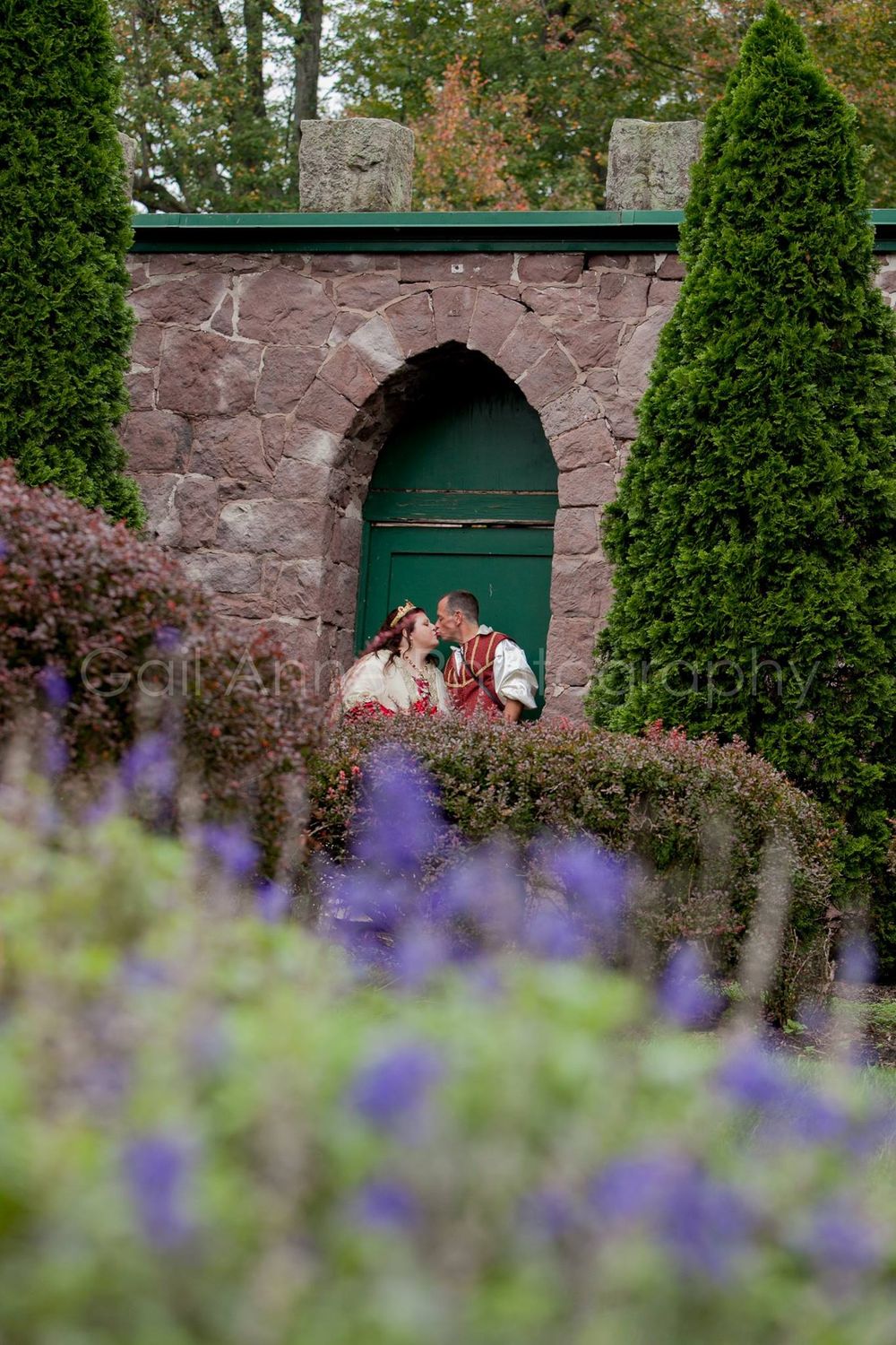 Couple in Renaissance attire sharing a kiss in a stone garden archway surrounded by purple flowers and evergreen hedges.