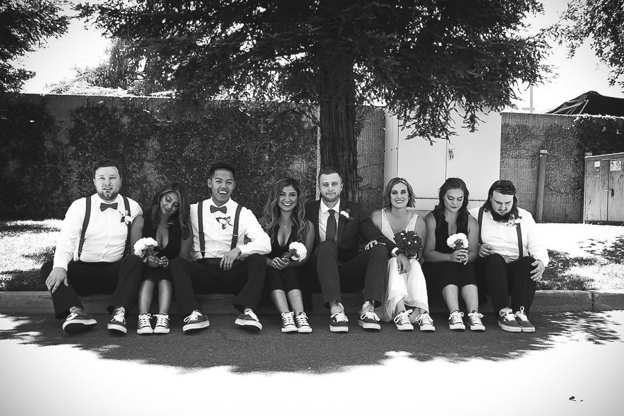 Black and white wedding party photo with bride, groom, and bridal party seated outdoors wearing suspenders and bow ties.