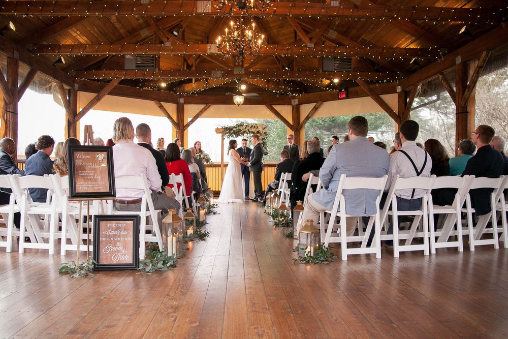 Elegant rustic wedding ceremony under a wooden pavilion with white chairs, floral decor, and warm string lights.