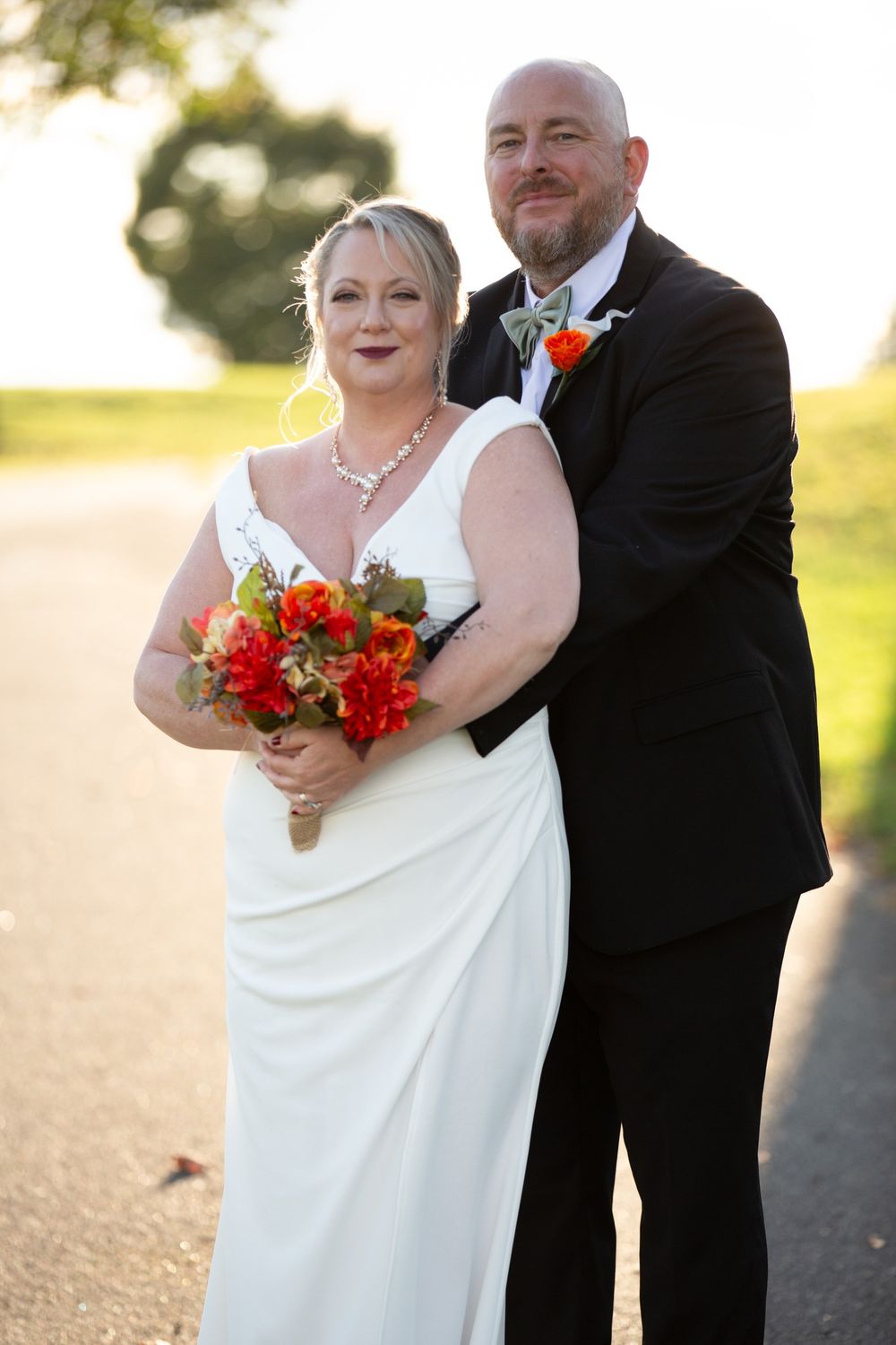 Bride in white gown holding red bouquet stands with bald groom in black tuxedo outdoors at golden hour.