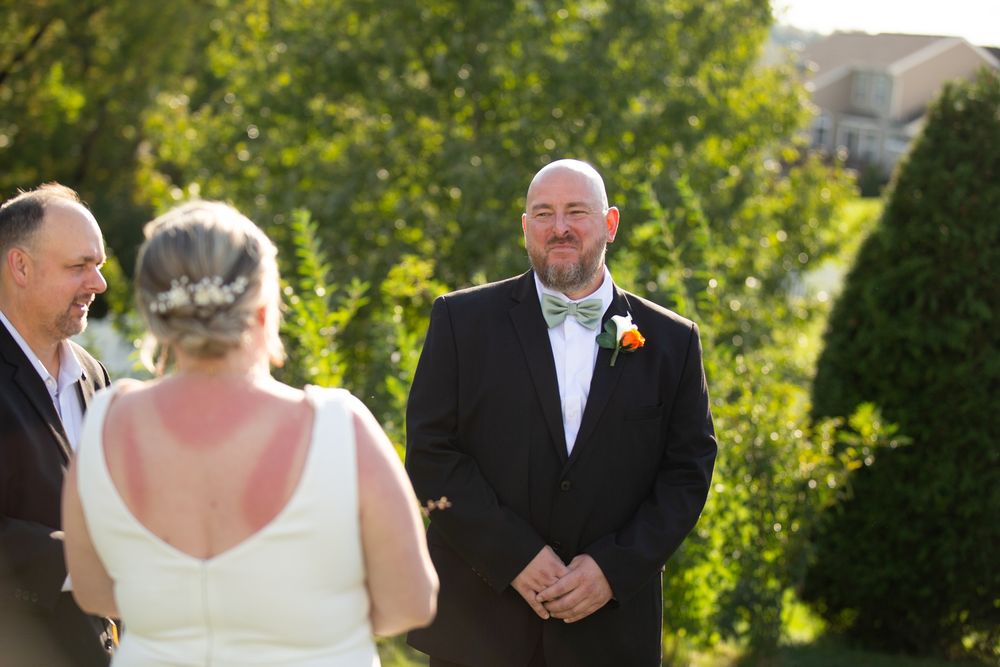 Groom smiling joyfully as bride walks toward him during outdoor wedding ceremony on sunny day.