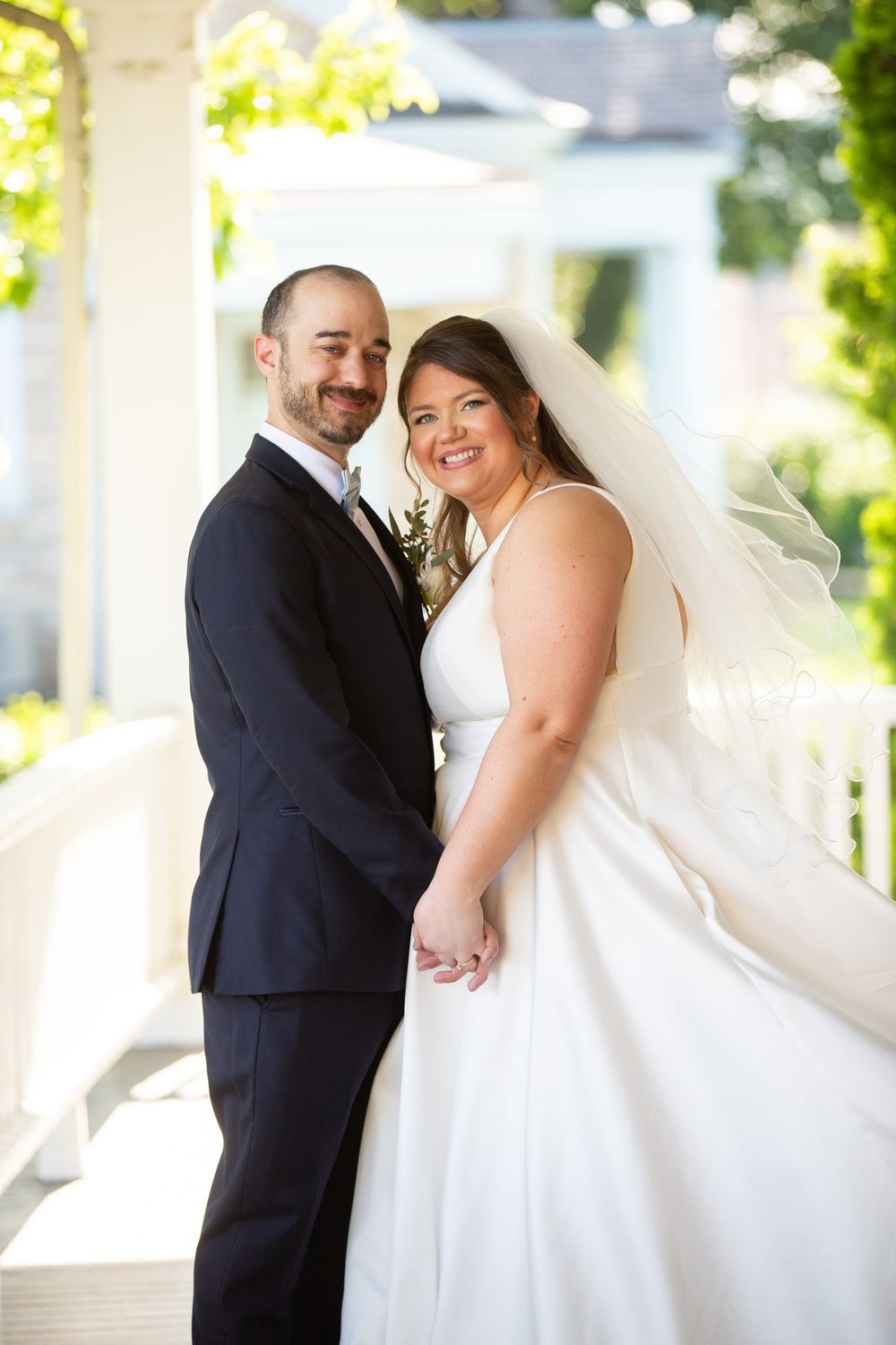 Smiling bride in white gown with veil holds hands with bearded groom in navy suit on sunlit porch.