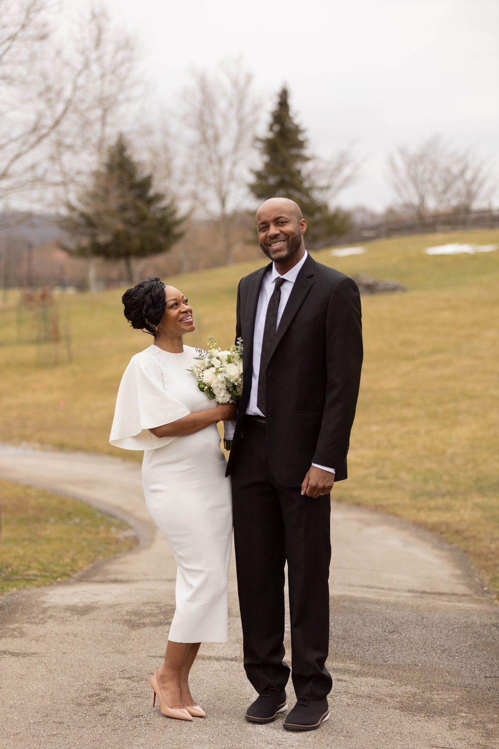 Happy couple in wedding attire standing on a path, bride in white midi dress holding bouquet, groom in black suit.